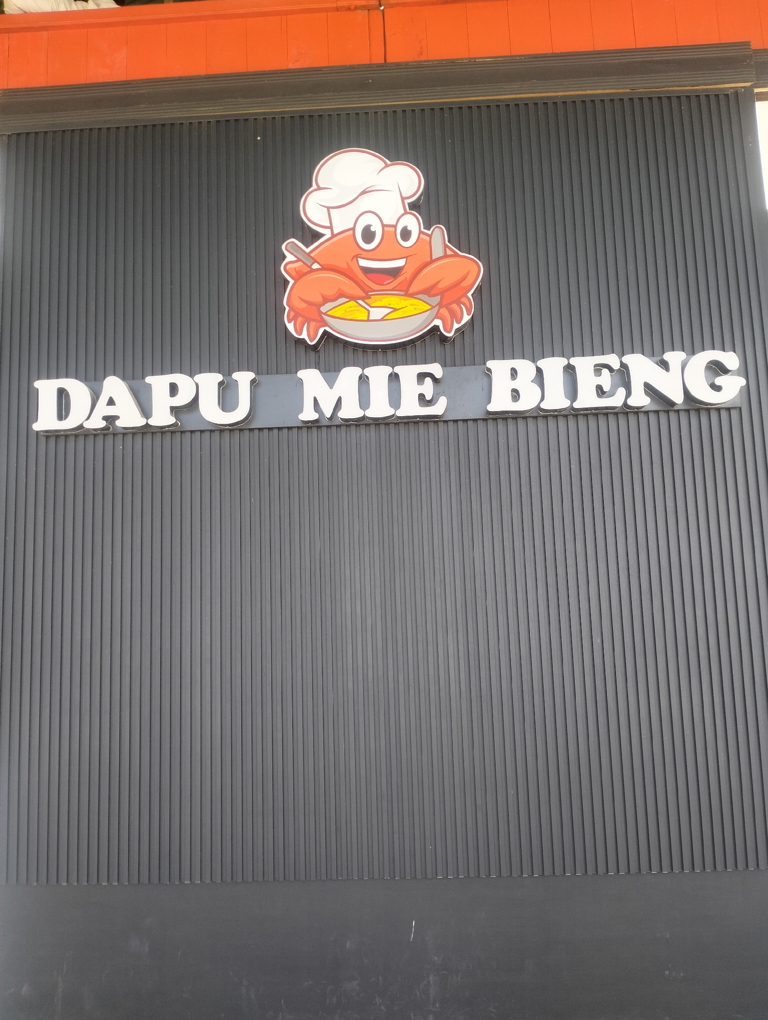 A sign with white block letters spelling "DAPU MIE BIENG" is mounted on a dark grey corrugated metal wall. Above the text is a cartoon illustration of a red crab wearing a chef's hat and holding a bowl of noodles with a fork. The crab has large googly eyes and a wide smile. Orange trim is visible above the grey wall. The scene appears to be an exterior storefront.