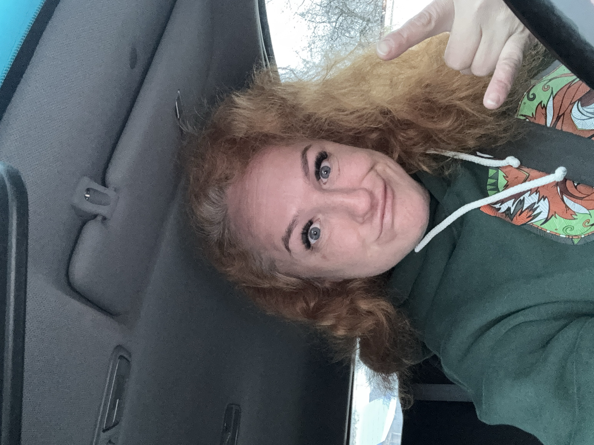 A woman with curly auburn hair and blue eyes sits in the driver's seat of a car. She wears a dark green hoodie and a seatbelt. Her right hand is raised, pointing towards her hair. The car's interior is visible, including the gray ceiling and a rearview mirror. The outside environment, visible through the window, appears to be wintery with bare trees. There is no evidence to indicate the city of Chaikovsky, Russia.