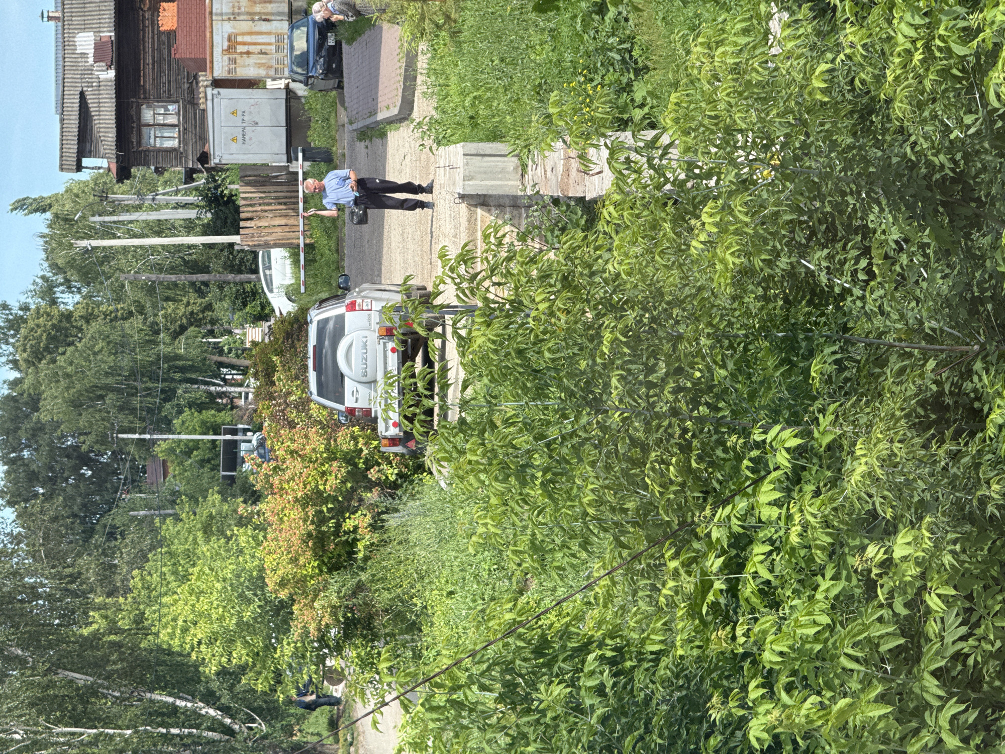 An outdoor scene in Votkinsk, Russia, shows a man holding a white barrier arm. He is standing on a paved surface next to a silver Suzuki SUV, with a black car parked to his right. A woman is standing near the black car, looking towards the man. In the background, a weathered wooden house stands beside shipping containers. Trees and lush green foliage dominate the foreground and surround the scene. The overall impression is a residential or semi-industrial area on a bright, clear day.