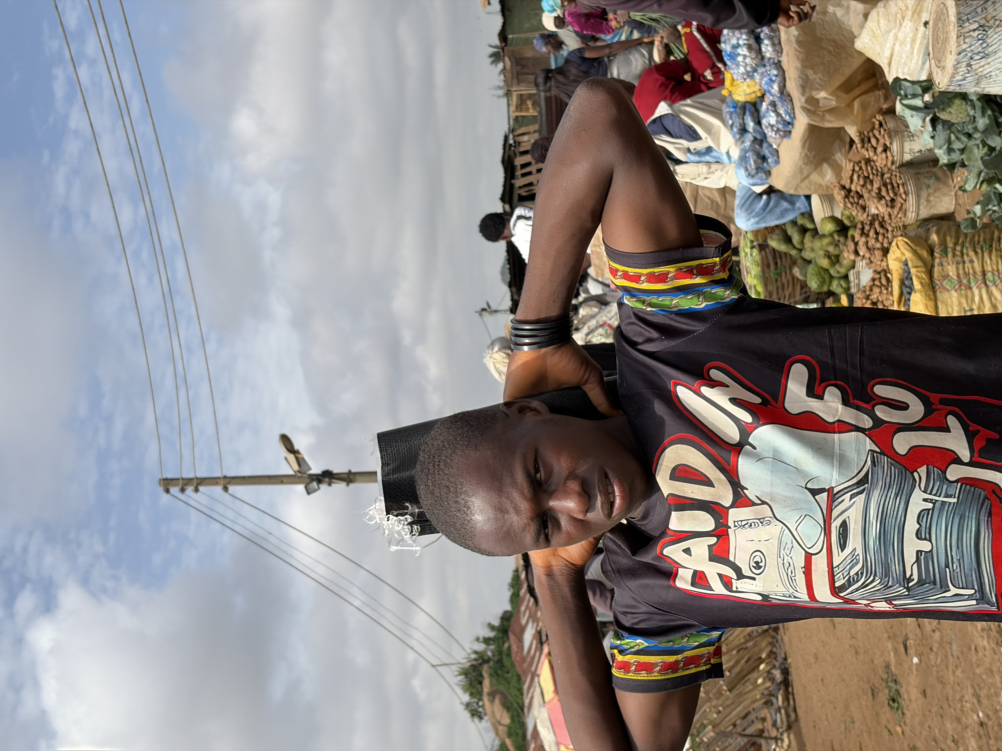 In Gura Top, Nigeria, a young Black male stands in a marketplace with his hands behind his head. He wears a black t-shirt with a graphic design of a hand holding stacks of money and the words "FAUD IN FLUX" printed in red and white. The man has short hair and is looking directly at the camera with a neutral expression. He is wearing black bracelets on his left wrist. The background of the image is blurred but shows a busy marketplace scene with stalls, produce, and other people. Overhead, power lines and a street light are visible against a cloudy sky.