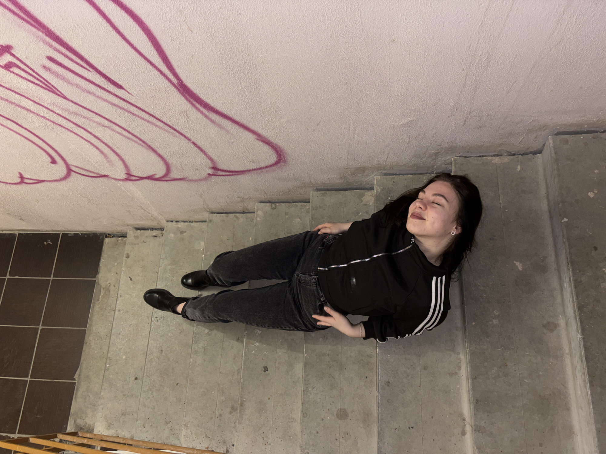 A young woman lies on a concrete staircase in what appears to be a stairwell. She is wearing black jeans, black boots, and a black Adidas jacket with white stripes on the sleeves. She is lying with her head near the upper part of the stairs and her feet near the bottom. Her eyes are closed, and she seems relaxed.
The stairwell has a concrete ceiling with pink graffiti. The stairs are also concrete. To the left of the stairs, there is a tiled floor. The location is likely in Perm, Russia.