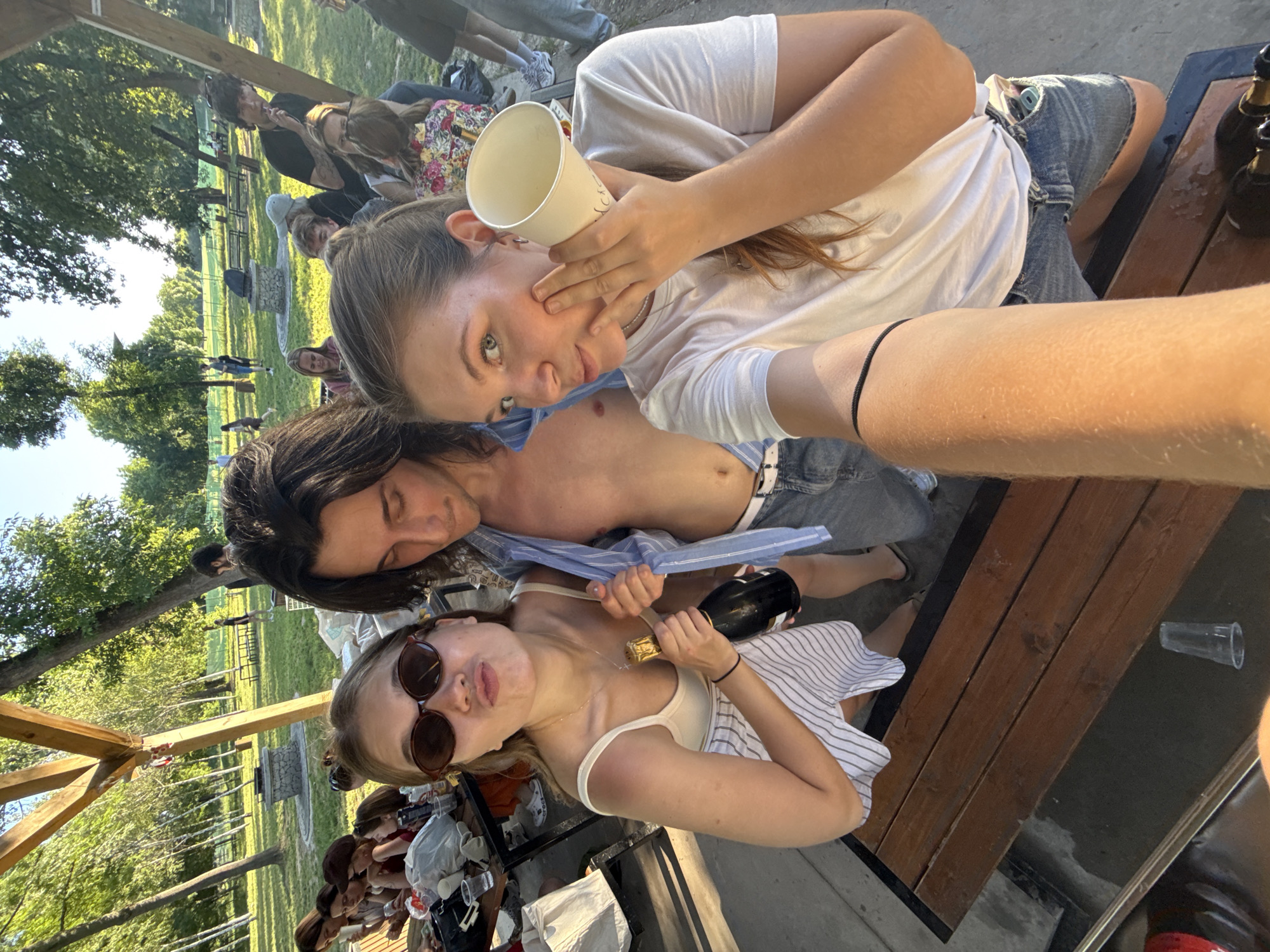 The image shows three young adults in an outdoor setting. The individual closest to the camera is wearing sunglasses and holding a bottle of champagne. Behind this person is another individual, who appears shirtless and wearing a striped shirt. The third individual is holding a paper cup and is wearing a white t-shirt. They are all gathered around a table. In the background, there are trees and other people. The photo was taken in the city of Cukarica Urban Municipality, Serbia.
