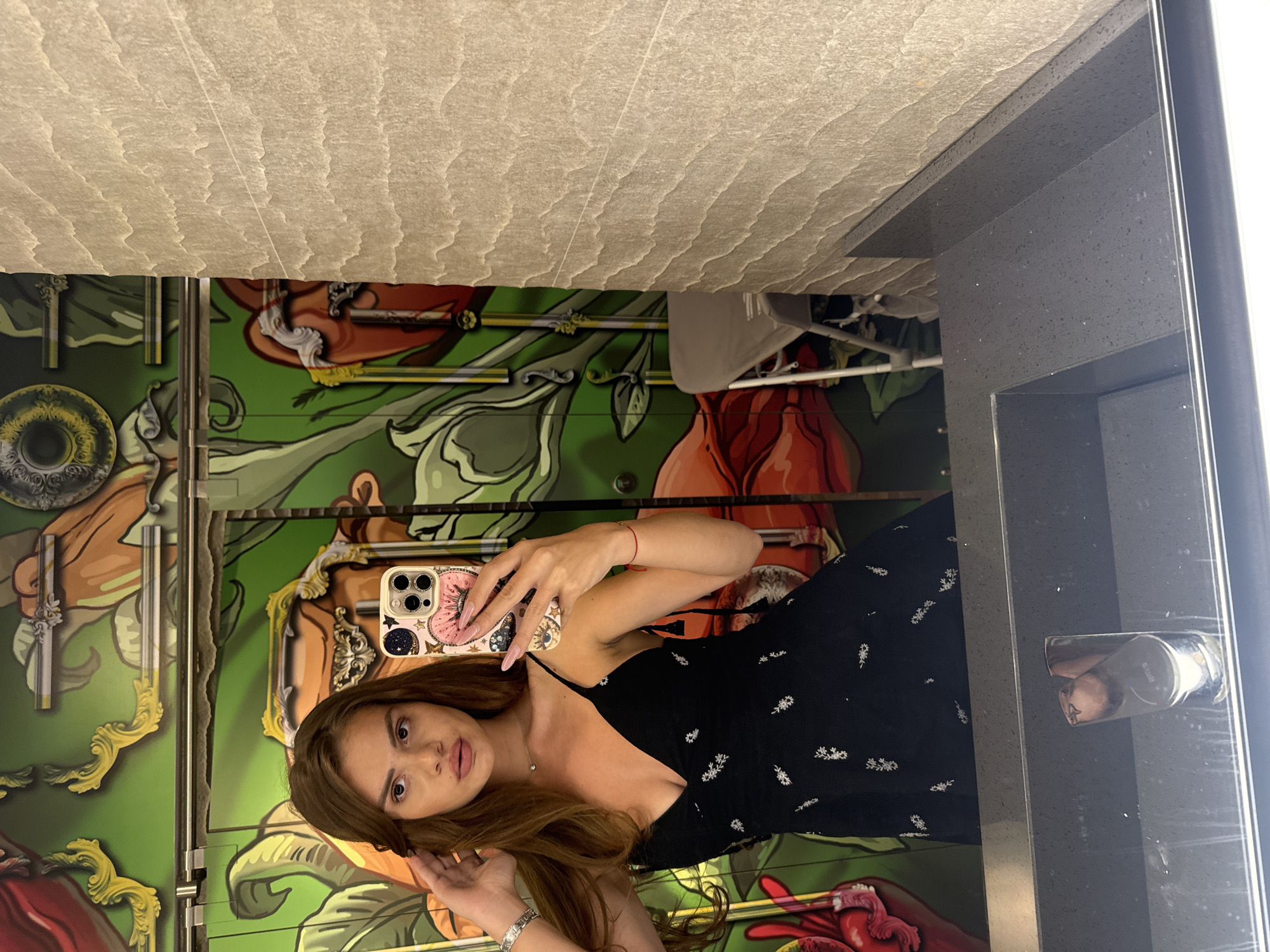 A young woman with long brown hair, wearing a black dress with a floral pattern and spaghetti straps, is taking a selfie in a bathroom. She is holding a phone with a pink case adorned with stars and a heart. Her left hand is touching her hair, and she is wearing a silver bracelet and a delicate necklace. The background features vibrant green graffiti with depictions of large flowers and abstract shapes. To the right of the graffiti, a textured beige wall is visible. A grey folding chair is partially visible in the reflection of the mirror. The sink area, made of dark speckled material, is in the foreground. The overall setting suggests an indoor location, possibly a restaurant, bar, or a public restroom with an artistic design, within the city of Plovdiv, Bulgaria.