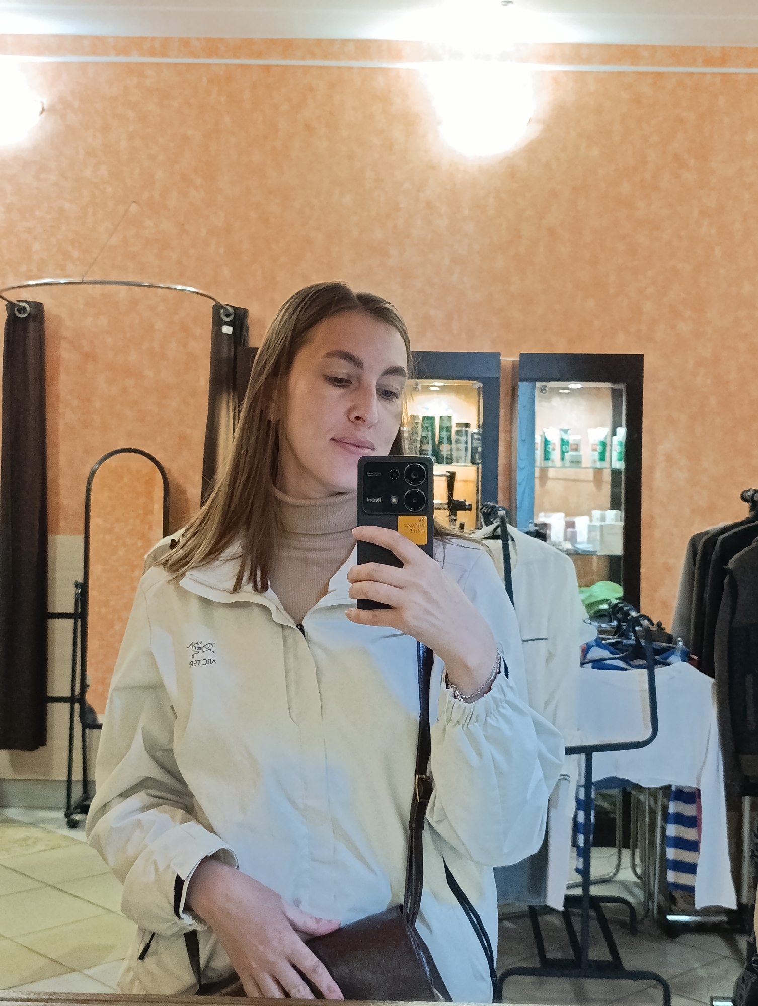 A young woman is taking a selfie in what appears to be a clothing store or boutique. She is wearing a light-colored jacket, a turtleneck sweater, and has a purse with a shoulder strap. The reflection shows clothing racks with garments, glass display cases filled with products, and orange textured walls. The location is Verkh-Sosnova, Russia.