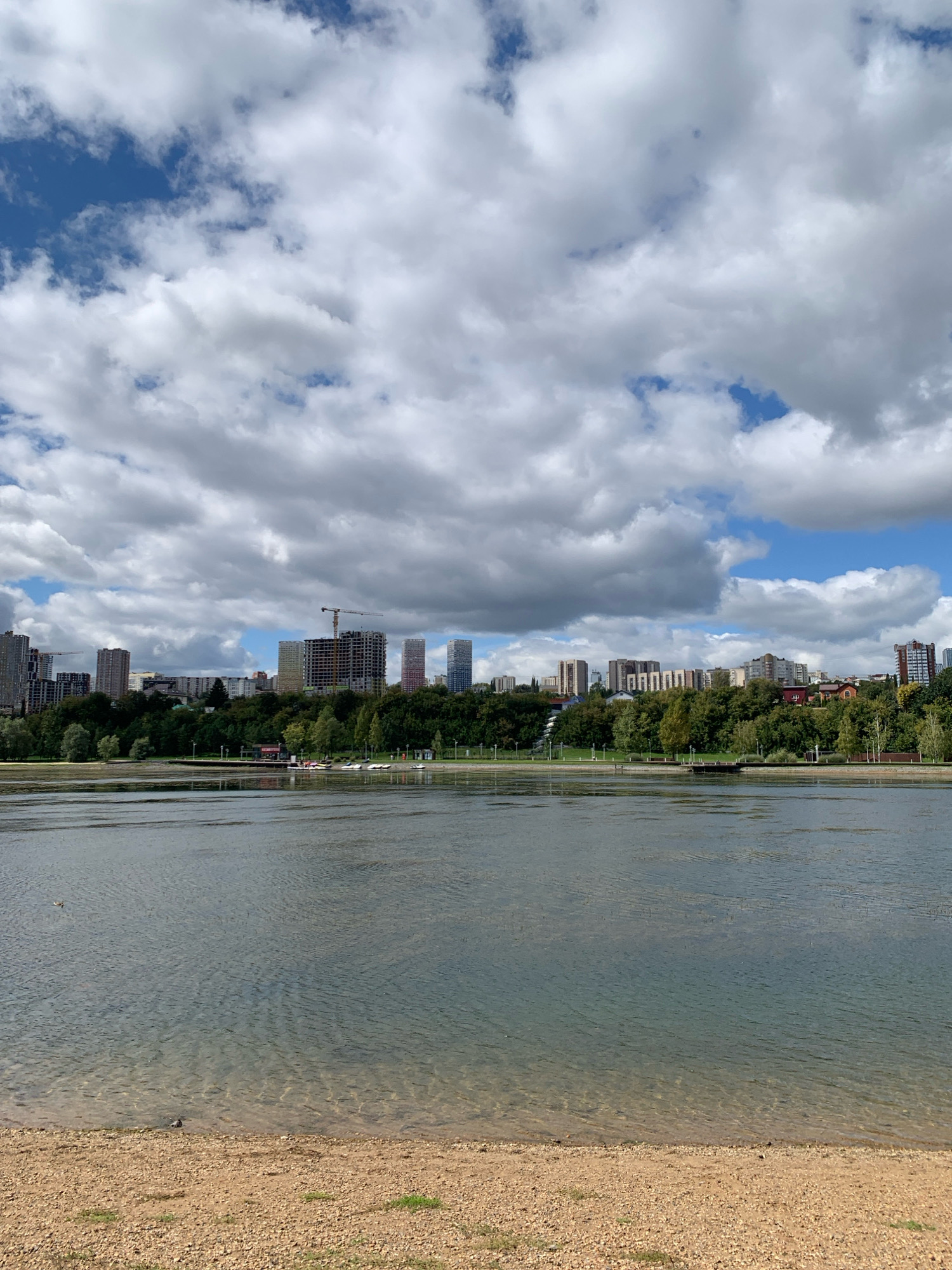 The image captures a scenic view of the Ufa city skyline across a body of water, likely a river or reservoir. In the foreground, a sandy embankment meets the calm, shallow water, with small pebbles and sparse green grass dotting the shore. The water exhibits a clear turquoise hue near the bank, gradually deepening in color. Two birds, one duck and one pigeon, are visible on the sandy shore.
The middle ground is dominated by a lush, green tree line that separates the water from the urban landscape. Beyond the trees, the Ufa skyline rises, featuring a mix of modern high-rise buildings under construction, some with cranes visible, and completed residential and commercial structures. The architecture suggests a contemporary urban development.
The sky above is dramatic, filled with a mixture of large, fluffy white clouds and patches of clear blue sky. The sunlight, filtered through the clouds, casts a soft illumination on the scene. The overall impression is one of a vibrant, growing city situated harmoniously with its natural environment.