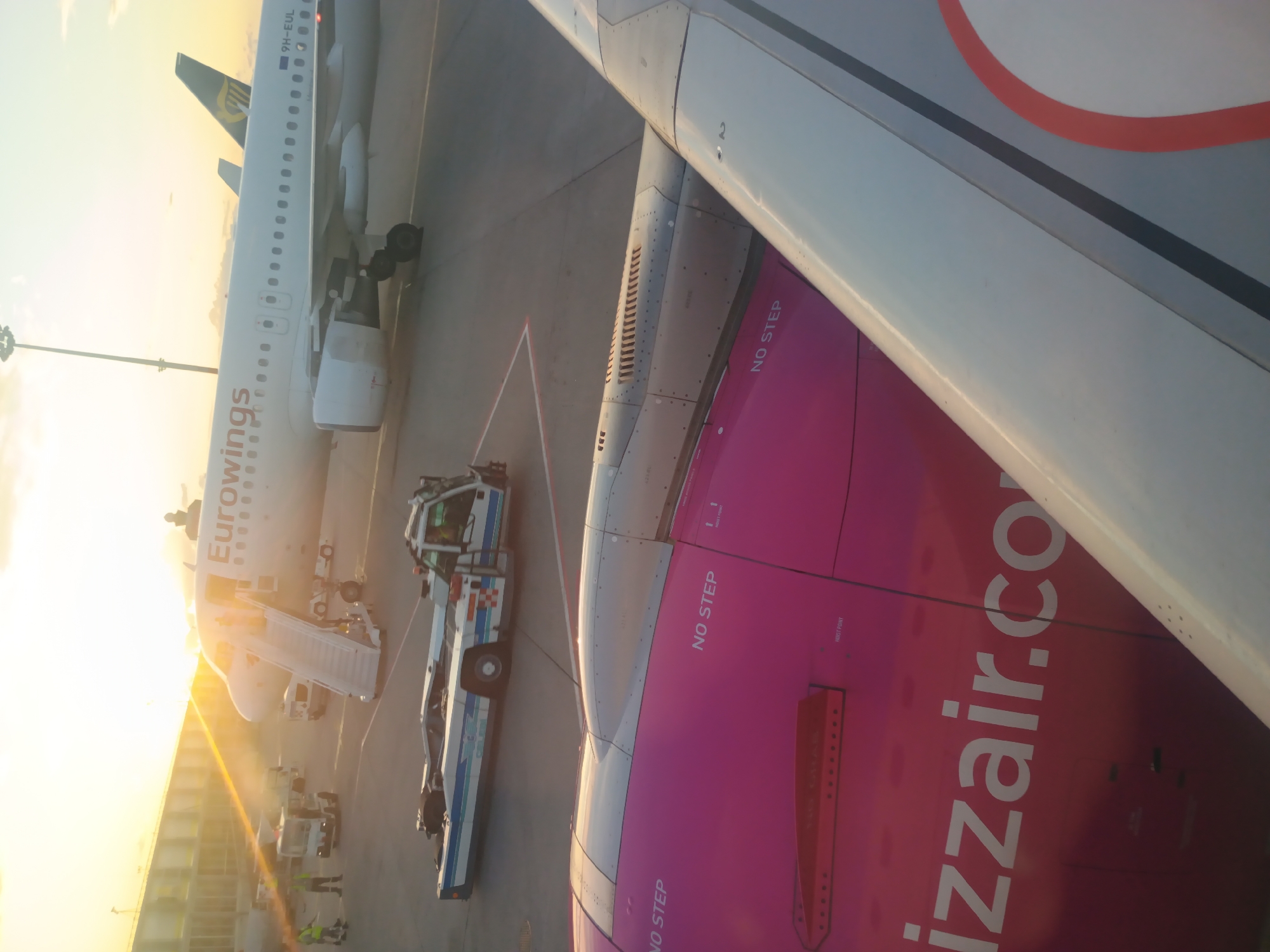 The image depicts an airport tarmac scene at sunset, likely at Budapest Ferenc Liszt International Airport, identified by the presence of Eurowings and Wizz Air aircraft, both of which have significant operations in Budapest. In the foreground, the wing and part of the fuselage of a Wizz Air plane are visible, featuring its distinctive pink livery and "wizzair.com" branding. The phrase "NO STEP" is clearly marked on the aircraft. In the mid-ground, a Eurowings aircraft is parked with a mobile passenger stair attached to its entrance. Various ground support vehicles, including a tug, are also present on the tarmac. The sky is illuminated by the warm glow of the setting sun, casting long shadows and creating a hazy atmosphere. The overall environment suggests a typical airport operational area, with the characteristic gray asphalt and painted markings.