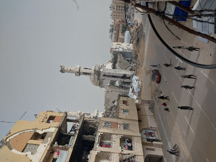 This image depicts a street scene in Khan Yunis, Palestinian Territory, characterized by extensive structural damage. In the left foreground, a multi-story building shows significant damage, with multiple floors partially collapsed, exposed interiors, broken windows, and debris; laundry hangs from balconies on its lower functional levels. Dominating the background is a heavily damaged mosque with a partially collapsed minaret and dome, surrounded by large piles of rubble. The paved street below features white line markings. Approximately 15-20 individuals are present on the street and sidewalks. A group of young males is dispersed across the road, some appearing to be walking or playing. On the left side of the street, an individual pushes another person in a wheelchair. A red sedan is parked on the right side of the street. Scattered debris, including corrugated metal, is visible near the damaged structures. The sky is overcast and hazy.