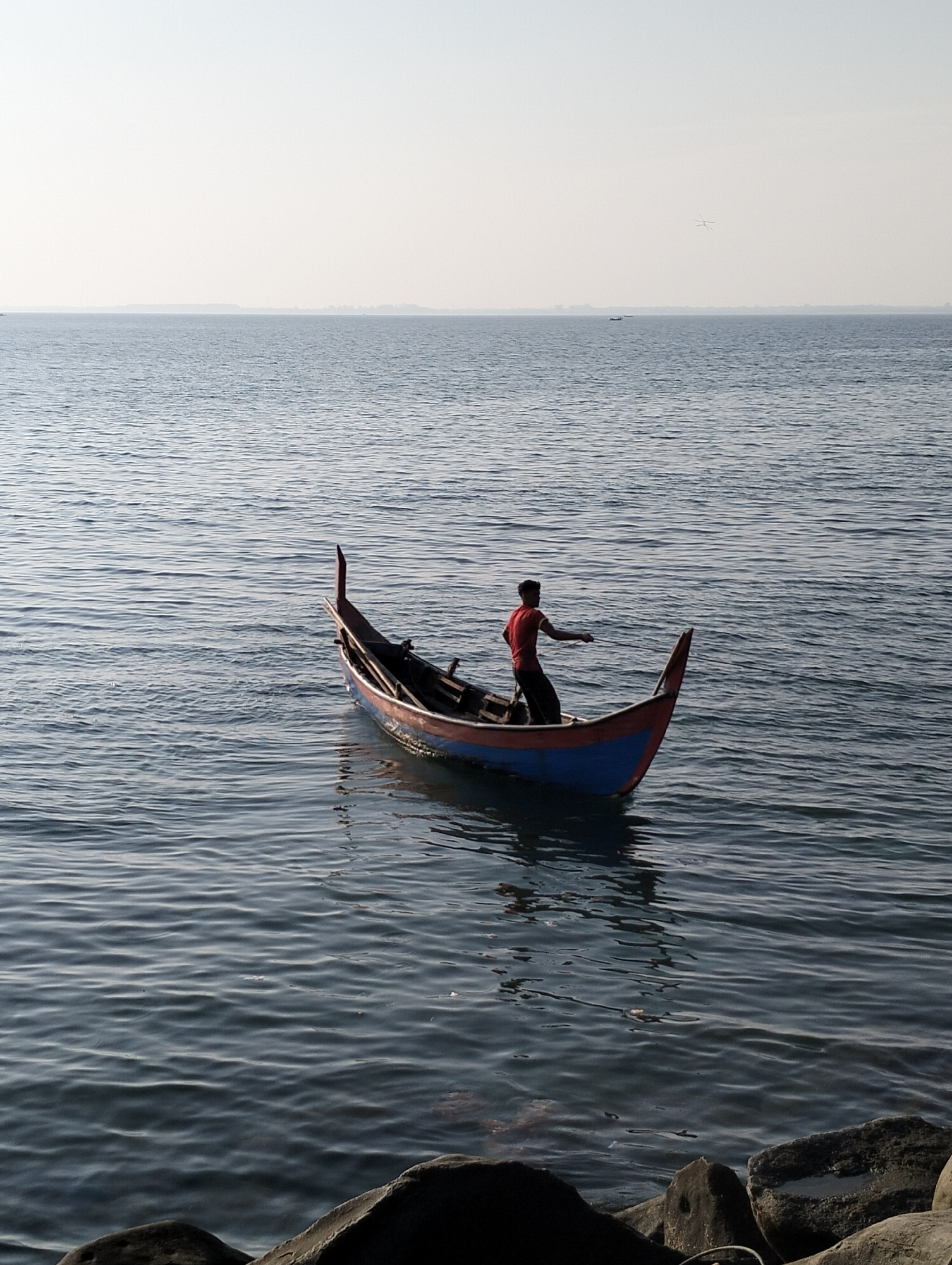 A medium-shot captures a solitary man aboard a blue and red boat, poised in the middle of the ocean. He wears a red shirt and dark pants, and his arm is outstretched, possibly engaged in fishing. The boat is centered in the frame. The sea extends from the foreground to the horizon, displaying gentle ripples. The sky is pale and overcast. The scene is in Lhokseumawe, Indonesia. In the foreground, partially obscured, are large, dark rocks. In the distance, at the horizon line, there are other boats and what appears to be a small plane or bird.