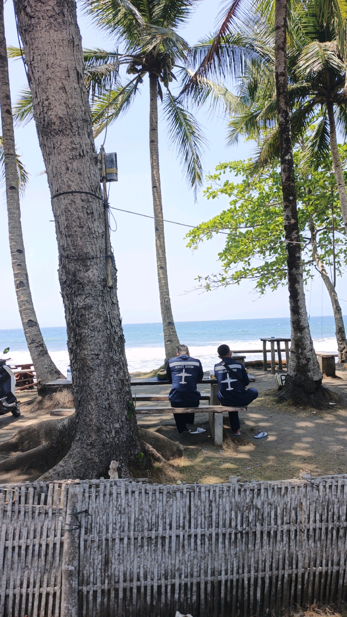 The image depicts a tropical beach scene in Desa Batukaras, Indonesia, with two individuals seated on a wooden bench facing the ocean. Both individuals are wearing dark blue jackets with white insignia, possibly representing an organization or team. The jackets have a white airplane graphic and text, which appears to read "RESOLUTE DEVOTION" on the left individual's jacket and "RESOLUTE DEVO..." on the right individual's jacket. They are positioned under the shade of mature coconut palm trees, with their backs to the camera.
The environment is characterized by lush vegetation, particularly tall palm trees with their fronds reaching towards a clear blue sky. The ground beneath the trees is sandy and covered with dry leaves and grass. In the foreground, a rustic fence made of vertical bamboo poles separates the beach area from the viewer.
Beyond the seating area, the ocean stretches out, with gentle waves breaking on the shore. The water appears a vibrant blue, indicating clear conditions. In the background, there are a few wooden structures, possibly tables or seating, and a motorcycle parked on the left side of the frame. The overall atmosphere suggests a relaxed and scenic coastal setting.