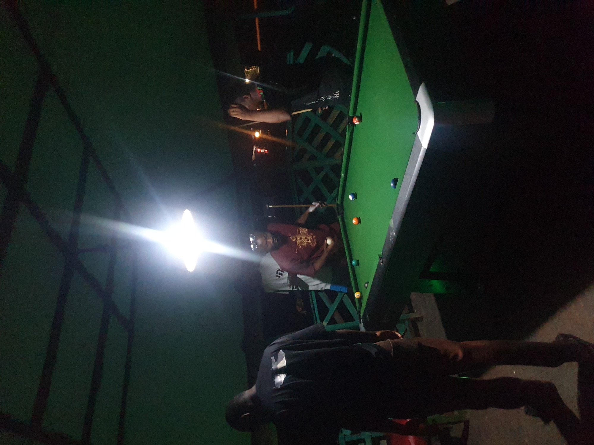 In Lokoja, Nigeria, at what appears to be an outdoor or semi-outdoor establishment, four individuals are engaged in a game of billiards under dim lighting, with a bright overhead lamp illuminating the pool table. One person, facing away from the camera and wearing a black t-shirt and brown pants, stands to the left of the table. Another man, in a maroon t-shirt, is positioned to the right, holding a cue stick and a white billiard ball. A third man in a white t-shirt stands behind the maroon-shirted player. The fourth individual is obscured, with only their silhouette visible. The pool table is green with colored balls scattered across its surface. The surroundings suggest a casual, possibly recreational setting, with a green roof structure overhead supported by dark beams. A green lattice-like railing is visible in the background. The overall atmosphere is dimly lit, with strong light emanating from the overhead lamp.