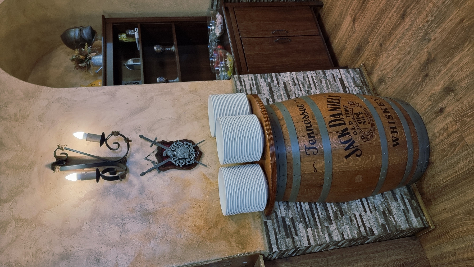 The image depicts an indoor scene, possibly a dining area or a bar, with a rustic and historical theme. In the foreground, a wooden barrel with "Jack Daniel's Old Time Old No.7 Brand Whiskey" prominently displayed is used as a pedestal for a stack of white plates. The barrel sits on a floor with wood-grain laminate planks. To the left of the barrel, a wall with a textured, cream-colored finish is adorned with a wrought-iron wall sconce featuring two electric candles. Below the sconce, a decorative crest with crossed swords is mounted. To the right of the barrel, a dark wooden cabinet with two doors is visible. On top of the cabinet, there are several glass bowls containing food items and a small watering can with dried flowers and a metal helmet. A dark wooden shelf unit with two shelves is situated behind the cabinet, displaying various items including glassware and decorative objects. The overall ambiance suggests a medieval or historical setting. The location in Besalú, Spain, is not visually confirmed by any explicit landmarks or signage within the image itself, but the architectural style, particularly the arched opening and the rustic decor, could be consistent with historical establishments in such a region. There are no people visible in the scene, and no active interactions are taking place. The lighting is warm and atmospheric, cast by the sconce and ambient room lighting.