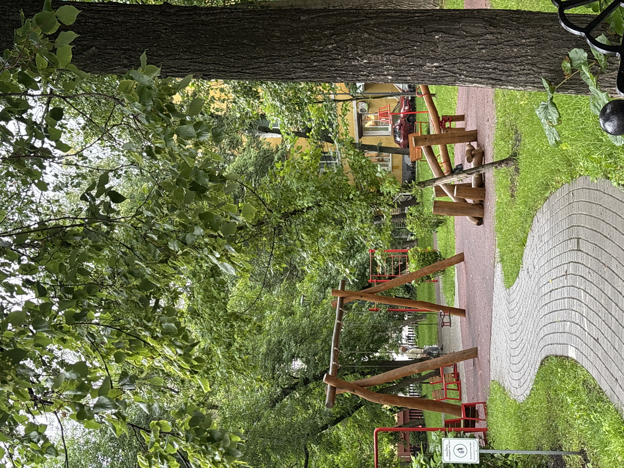The image depicts an outdoor playground in a city. In the foreground, a curved paved path made of light-colored bricks winds through lush green grass. To the left of the path, a white sign with a symbol of a trash can and litter is visible. Further to the left, a red bench is situated next to a playground structure.
Dominating the midground is a wooden swing set with two swings. Red metal playground equipment is also present in the background. To the right of the swing set, a wooden seesaw-like structure is visible. A large tree with dark bark stands prominently on the right side of the image. Behind the playground, a yellow building with multiple windows can be seen, and a red car is parked near it.
The environment is filled with dense green foliage, suggesting a park or courtyard setting within a city. The lighting indicates it is daytime, possibly overcast. The scene is devoid of any overt action or interaction among people. There is a single person visible in the background, standing near the red benches, appearing to be walking away from the camera. The overall impression is one of a peaceful, slightly weathered urban playground.