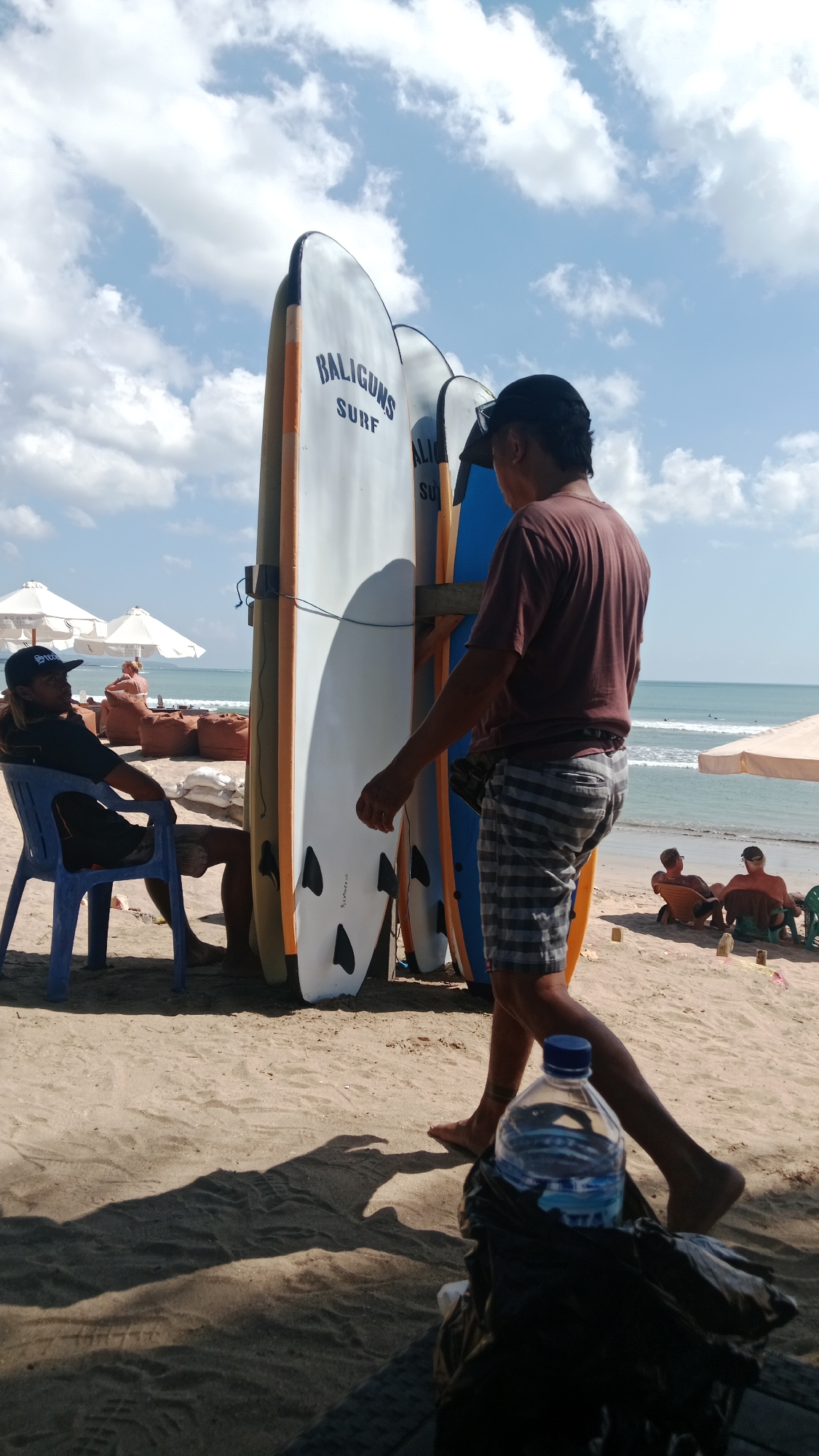 The image captures a sun-drenched beach scene in Kuta, Indonesia, likely near a surf school given the prominent display of surfboards. In the foreground, a person wearing a maroon t-shirt and checkered shorts walks away from the camera, casting a long shadow on the sandy ground. To the left, another individual sits in a blue plastic chair, wearing a black t-shirt and a black baseball cap, looking towards the ocean. Several surfboards, some white and others blue, are propped upright on a wooden stand. In the background, the turquoise ocean stretches out to the horizon, with a few distant surfers visible. White umbrellas and scattered beanbag chairs suggest a relaxed beachside atmosphere. The sky is a bright blue, dotted with fluffy white clouds. The scene evokes a typical day at a popular Indonesian beach destination.