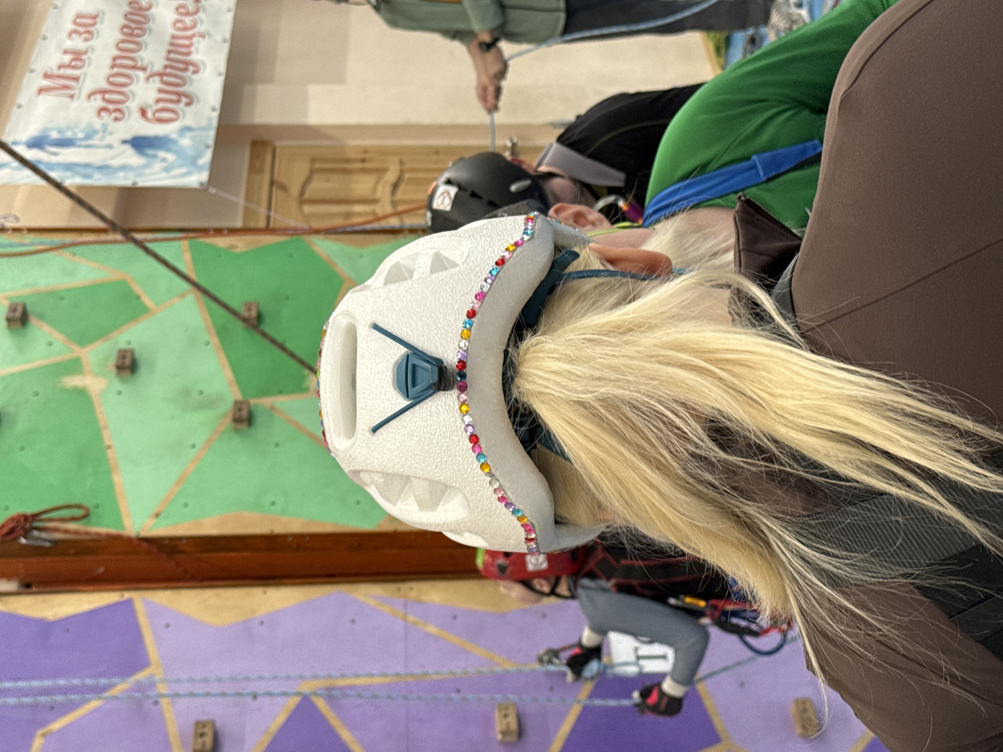 A climbing gym in Unknown, Unknown, features a young woman wearing a white helmet adorned with colorful rhinestones. She has long blonde hair, and is dressed in climbing gear, secured by ropes, and facing a climbing wall. The wall is sectioned into green and purple segments with various handholds. Additional people are visible, one wearing a black helmet, and another with a watch. A banner hangs in the background with text in an unidentified language.