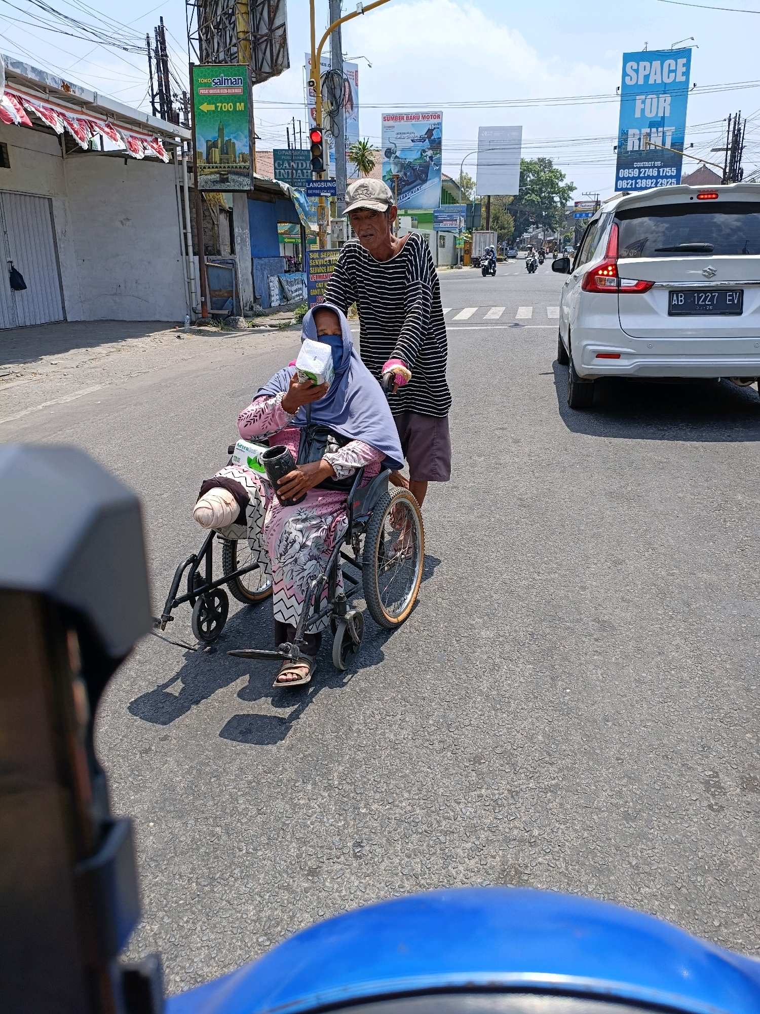 A man pushes a woman in a wheelchair on a street in Maguwoharjo, Indonesia. The woman is wearing a blue hijab and a pink patterned dress, and has a prosthetic leg. She is holding a box of tissues. The man is wearing a striped shirt and a hat. A white SUV is stopped in the background. The street is paved and there are buildings and billboards on either side. The sky is blue and there are some clouds.