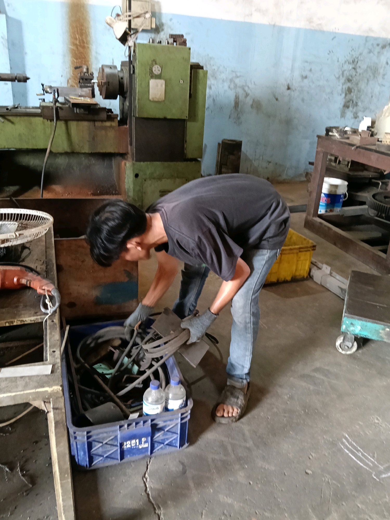A male is seen in a workshop in Belimbing, Indonesia, wearing a black t-shirt, blue jeans, and sandals. He is wearing grey work gloves and appears to be sorting through a blue plastic crate filled with metal parts. The crate contains various metal components, possibly scrap metal or machine parts. Two plastic bottles are visible inside the crate. In the background, there is a large green industrial machine, likely a lathe, and other workshop equipment, including a fan and a toolbox. The floor is concrete, and the walls are painted blue and are marked with dirt and smudges. The lighting suggests an indoor environment, possibly with natural light from a window or overhead fixtures.
