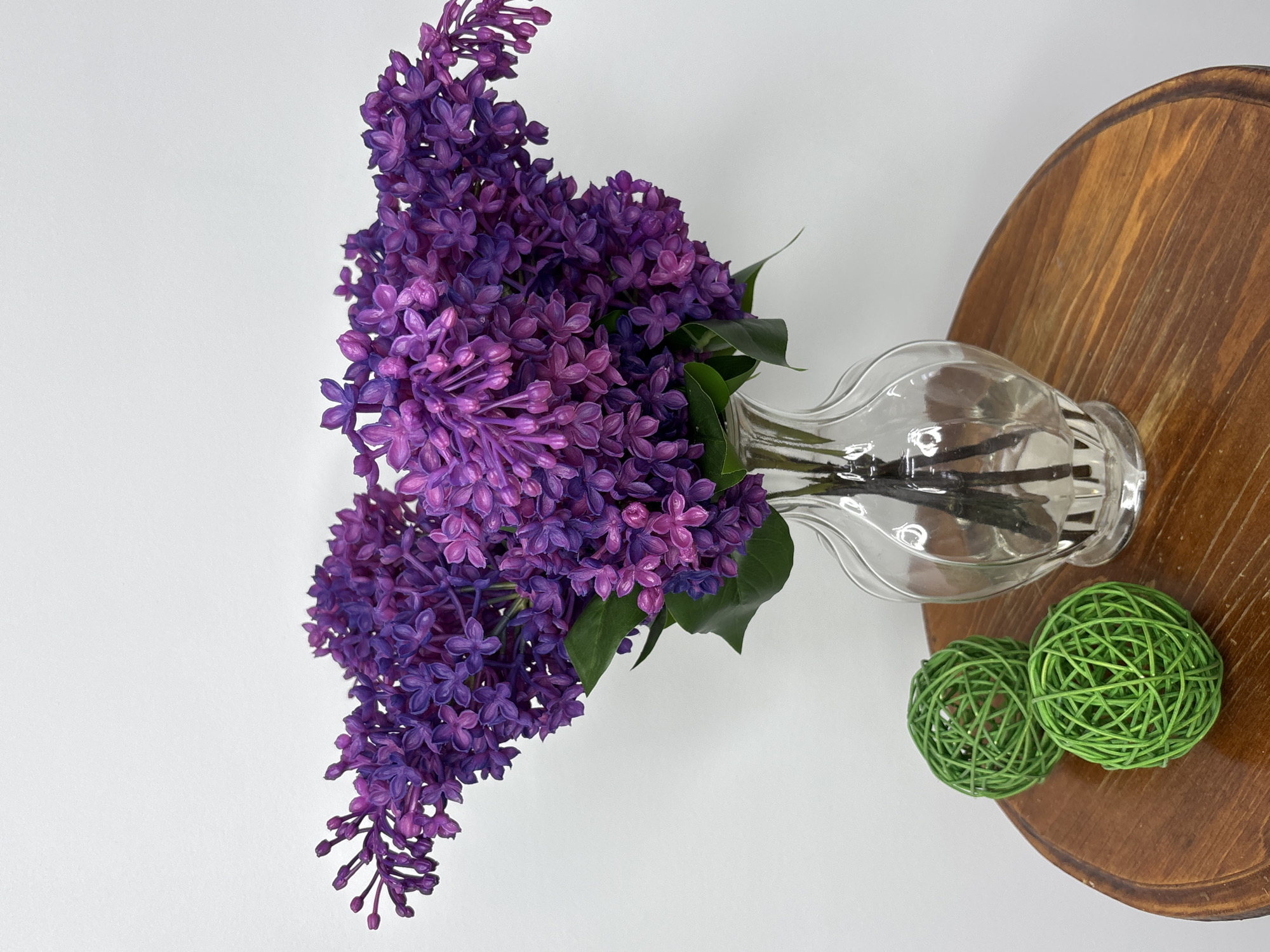 A photograph taken from above displays a bouquet of purple lilacs in a clear glass vase, placed on a wooden circular surface alongside two green decorative balls, against a white background. The lilacs are densely packed, showcasing various shades of purple. The clear glass vase reveals its internal structure and stems. The wooden surface features a pronounced wood grain pattern. The two green balls are made of interwoven strands. The background is plain white. There is no indication of this photograph being taken in Chaikovsky, Russia.