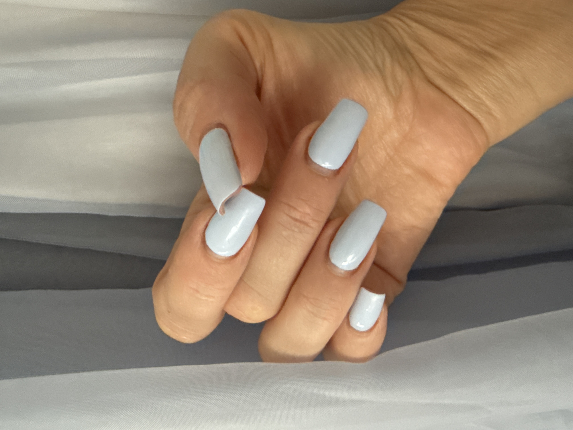 A close-up, slightly angled shot shows a person's right hand with light blue, coffin-shaped acrylic nails against a background of light gray and white sheer curtains. The hand is positioned in the foreground, with the fingers curved and the thumb slightly visible at the bottom right. The nail polish is glossy and uniform in color. The curtains in the background are draped and softly lit, creating vertical lines and a slightly diffused effect. No specific location in Kadıköy, Turkey is identifiable in the image.