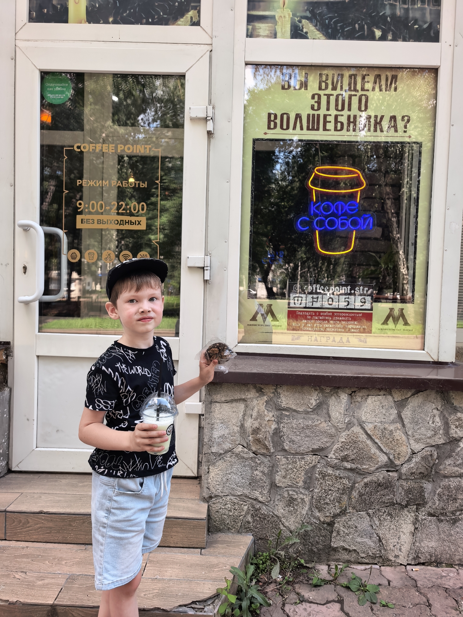 This media file captures a young boy standing outside a "Coffee Point" establishment in Sterlitamak, Russia. The boy, wearing a black t-shirt with a white graphic print and a black baseball cap, holds a cup with a white beverage and a pastry in a plastic wrapper. The background features the entrance to the coffee shop, with glass doors displaying business hours and promotional text in Russian. Notably, a neon sign in the window reads "Coffee To Go" and a large banner asks, "Have you seen this magician?". The scene appears to be set during the daytime, with natural light illuminating the exterior. The overall mood seems casual and lighthearted.