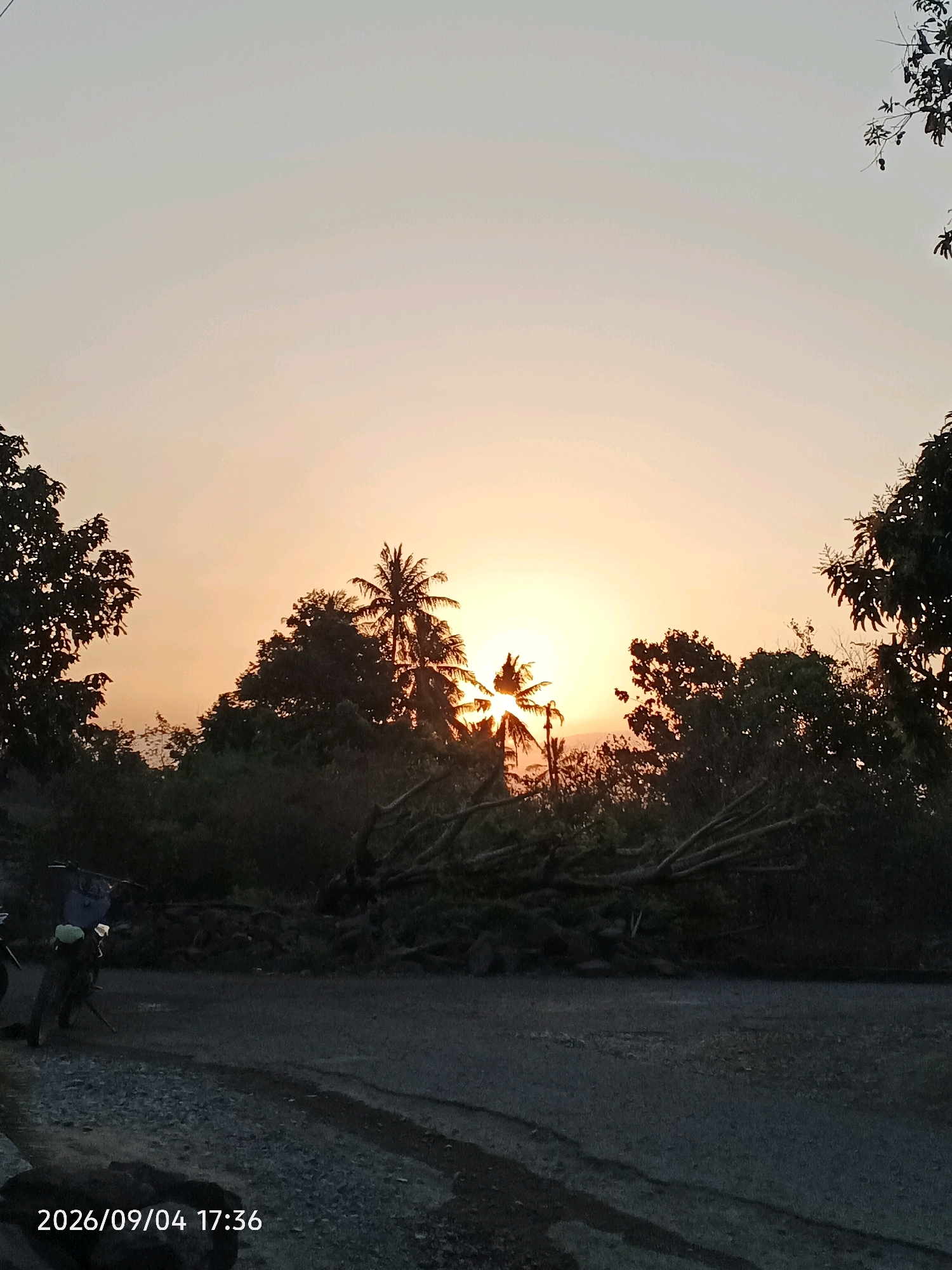 The image captures a sunset in Bulu Cenrana, Indonesia, with the sun partially obscured by trees. The sky transitions from a pale yellow near the horizon to a light gray at the top. Silhouetted trees, including a prominent palm tree, frame the sun. In the foreground, a dirt road is visible with a motorcycle parked on the left side. A person, dressed in dark clothing and wearing a backpack, is seen standing next to the motorcycle. The time stamp "2026/09/04 17:36" is present in the bottom left corner.