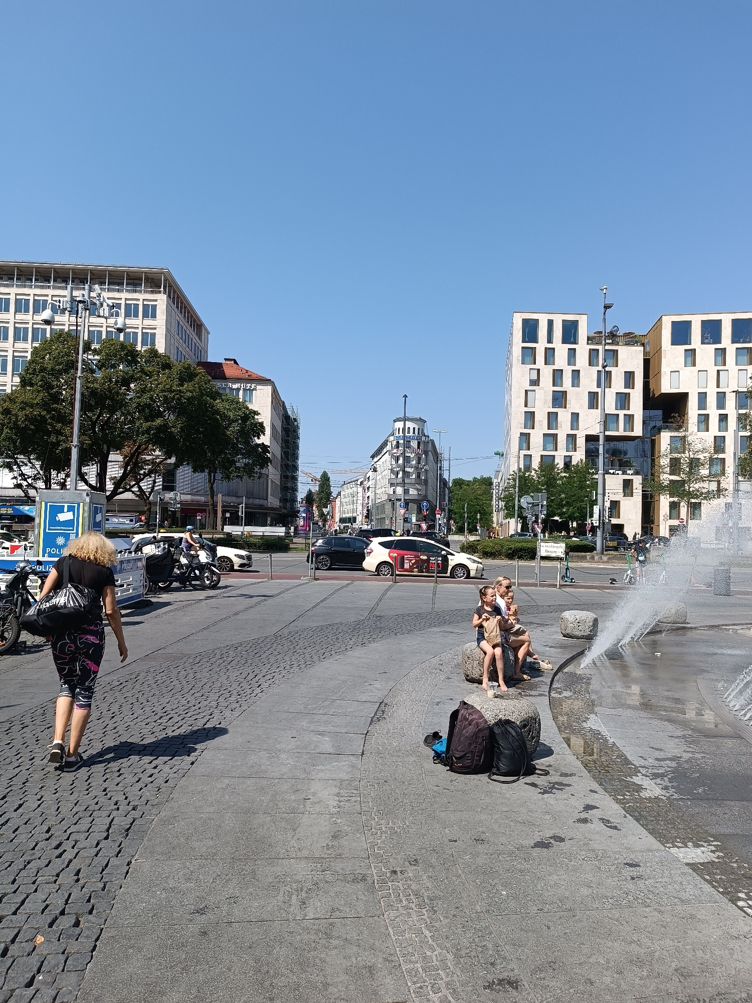 The image depicts a sunny day in a pedestrian area of Munich, Germany, likely a public square with cobblestone and paved sections. In the foreground, a woman with blonde hair walks away from the camera, carrying a large black bag. She is wearing patterned leggings and a black top. To her left, bicycles are parked. Further into the scene, several people are seated on large stones near a fountain, with water spraying. One individual, possibly an adult, is with two children, and they appear to be eating or sharing food. In the background, there are modern buildings with many windows, as well as older architectural styles. Cars and what appears to be a police vehicle are visible on a road or thoroughfare in the mid-ground. Trees and streetlights are also present, contributing to the urban setting. The sky is clear and blue.
