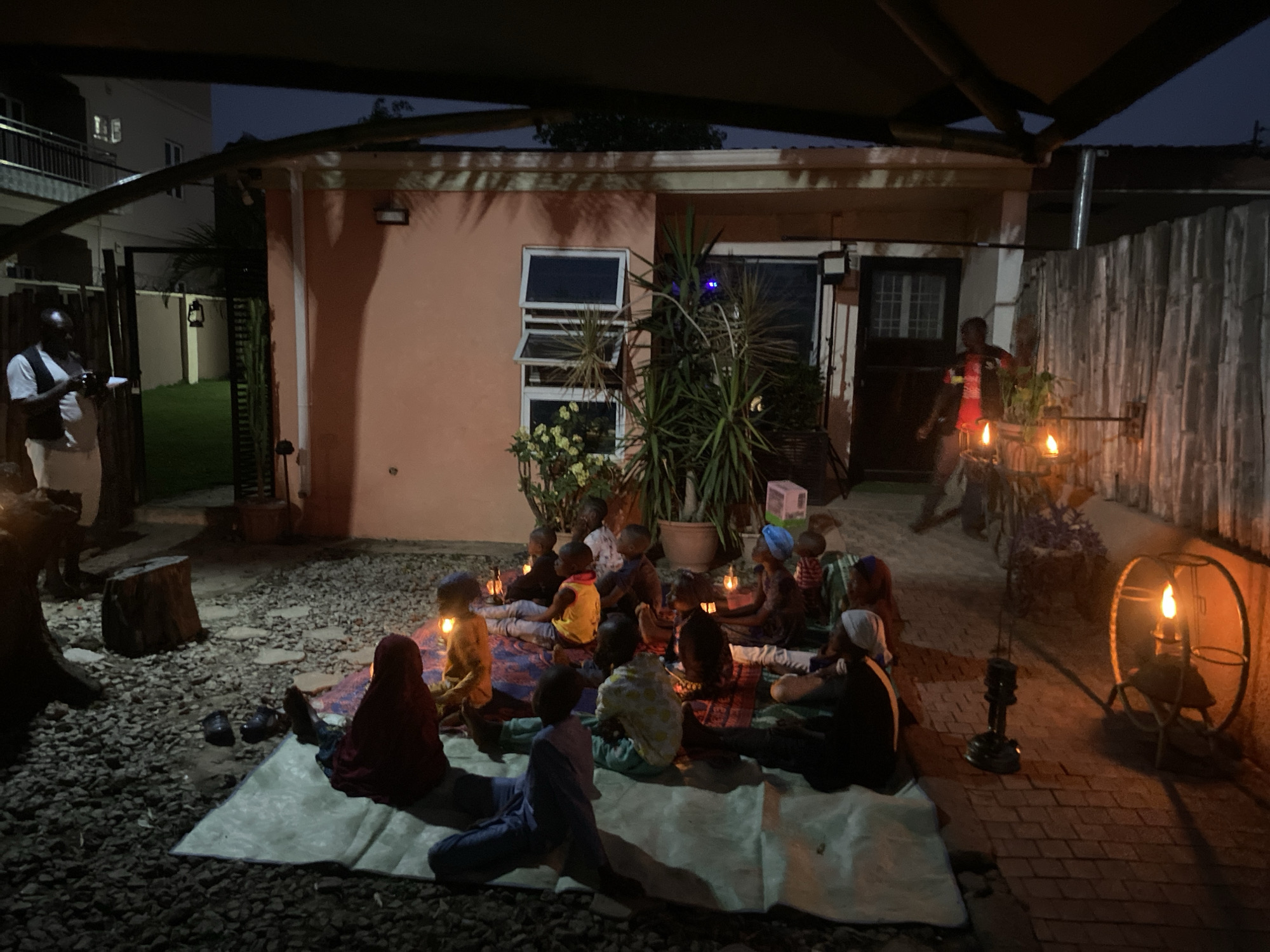 In Barden Yana, Nigeria, at night, a group of children are sitting on mats and blankets in an outdoor courtyard area, likely engaged in some activity. They are gathered around a central area, and there are individual lanterns placed beside some of them, providing light. The scene is set with a building behind them that has a light-colored exterior with windows and a door. Adjacent to the building is a wooden fence with lanterns or candles attached. Several adults are also visible, including one standing on the left of the frame holding a device, possibly a camera. The overall atmosphere is illuminated by the lanterns and the setting of the night sky. The surface of the yard is covered in loose stones and some brick tiling toward the right. A large potted plant is positioned near the building.