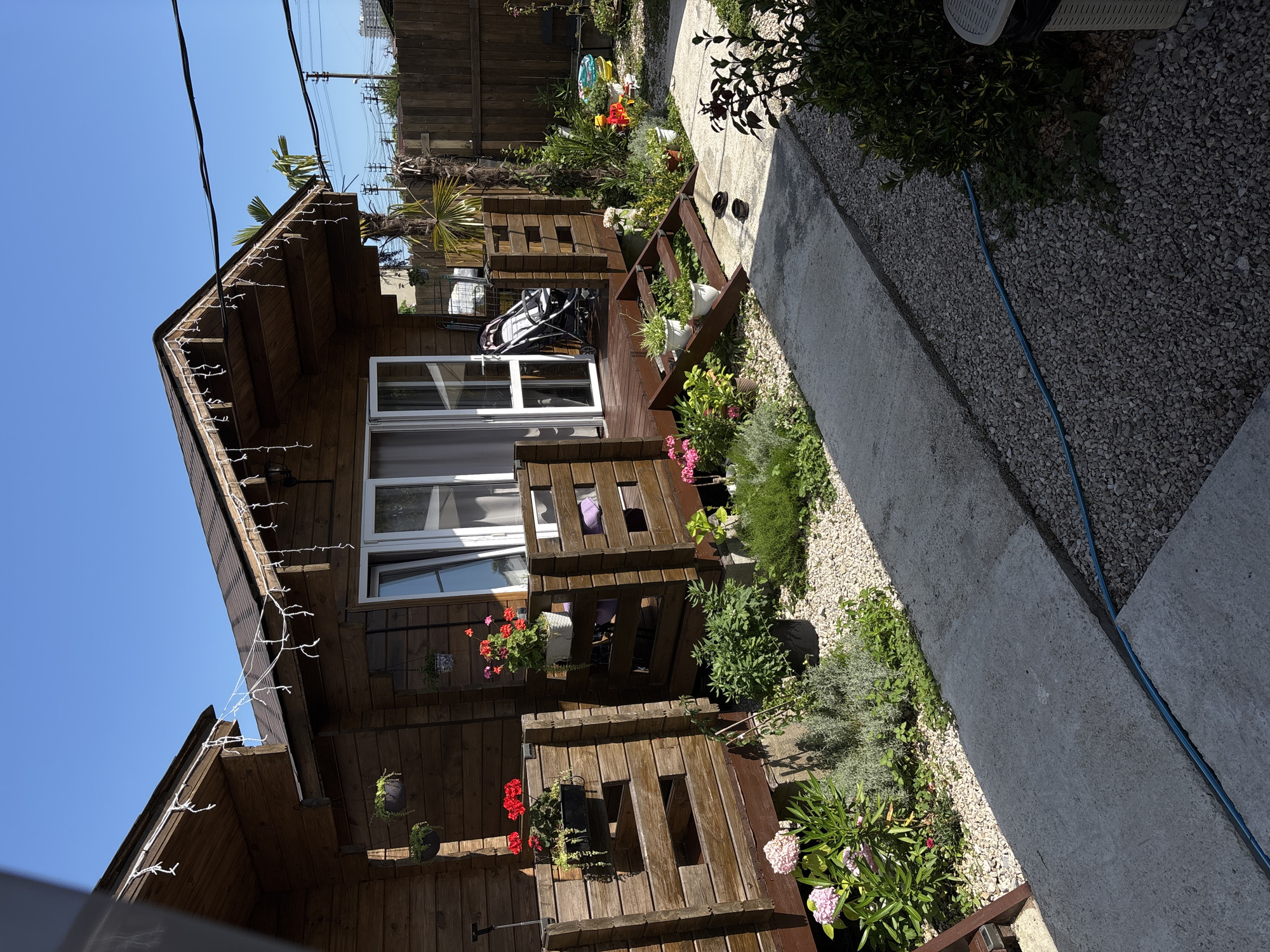 The image depicts an outdoor scene of a rustic wooden house with a garden and a concrete path. The house appears to be a small, two-story structure with wooden siding and a sloped roof. String lights are draped along the eaves of the roof. There are several windows and balconies on the house, some of which are decorated with potted plants.
The garden is filled with a variety of plants and flowers, including pink and red flowers, green bushes, and some palm trees. The ground is covered with gravel and some patches of grass. A concrete path runs along the side of the garden, and a blue hose is visible on the gravel.
In the foreground, there are some children's toys, including a red toy car and a blue and yellow toy. There is also a stroller visible on a wooden deck. The scene appears to be set during the daytime, with bright sunlight and a clear blue sky, suggesting fair weather. The overall impression is one of a cozy and lived-in outdoor space, possibly a backyard or a patio area. The location is identified as Gudauta, Abkhazia. No people are visible in the image.