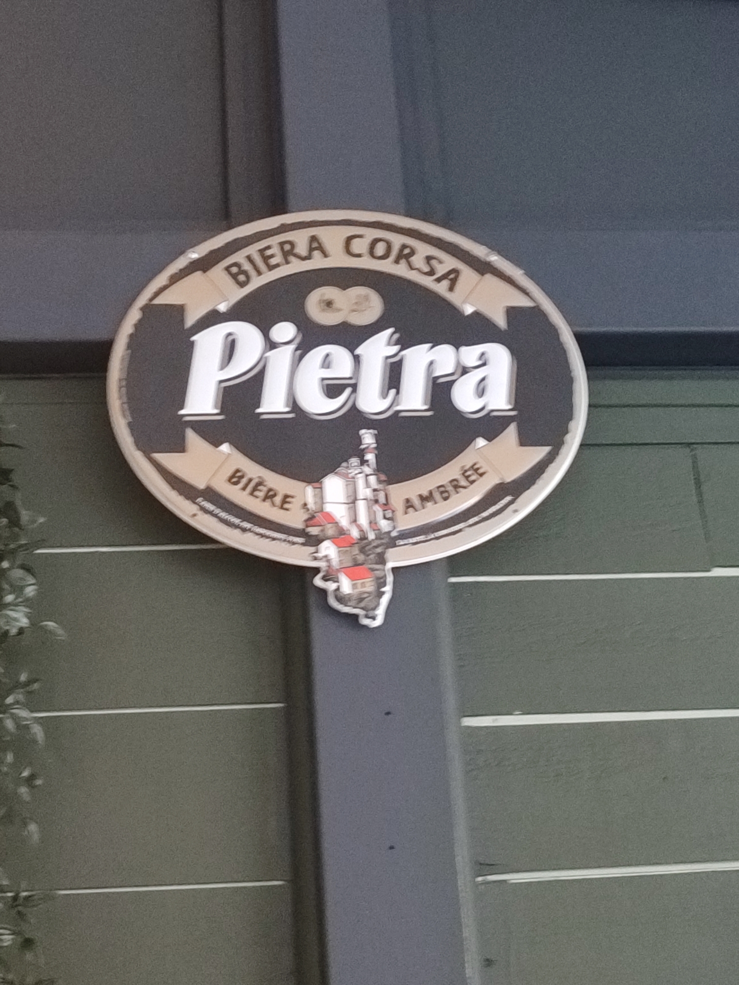 A round, dark-colored sign with a beige border is displayed on a dark gray pole. The sign features white text that reads "Pietra" prominently in the center. Above "Pietra," curved text says "BIERA CORSA." Below "Pietra," on two beige banners, are the words "BIERE" on the left and "AMBRÉE" on the right. A small, stylized illustration of a building complex with red roofs is attached to the bottom of the sign, overlapping the "BIERE" banner. The background of the image shows a dark green, horizontally paneled wall. The sign is outdoors, mounted on a pole in what appears to be a public space or establishment in Hyères, France, though the specific location within Hyères cannot be determined from the image alone. No people are visible in the frame.