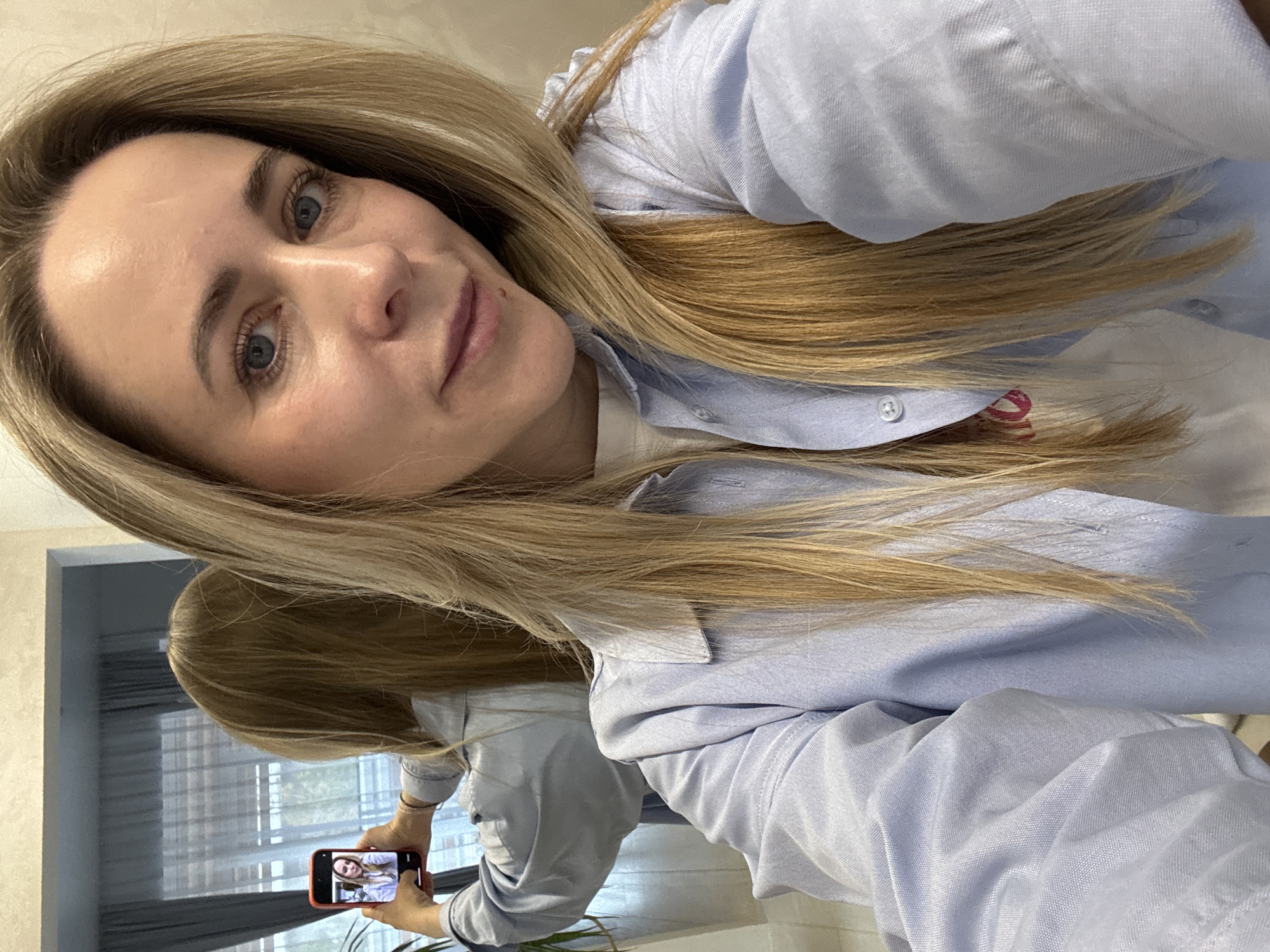 A woman with blonde hair and blue eyes is taking a selfie with her phone in a mirror. She is wearing a light blue button-down shirt over a white top. The reflection in the mirror shows the woman holding her phone, taking the picture. The background includes a window with white curtains and a hint of greenery outside. The photo was likely taken indoors, as there are interior elements visible. There is no information indicating the photo was taken in Chaikovsky, Russia.