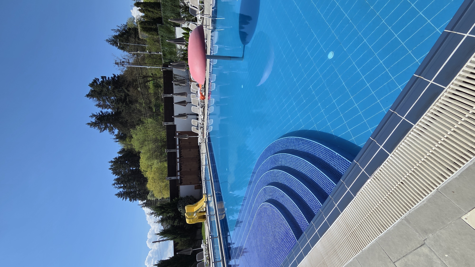 The image captures a vibrant outdoor swimming pool scene under a bright, clear sky. The main subject is a large, rectangular swimming pool filled with sparkling blue water, reflecting the clear sky above. The pool floor is visible through the clear water, revealing a distinct pattern of blue square tiles. At one end of the pool, elegant curved steps, also tiled in blue mosaic, descend into the water. A light-colored drainage grate runs along the edge of the pool, bordered by darker blue or black tiles.
Along one side of the pool, numerous sun loungers are neatly arranged, mostly white, with some accompanied by small tables and dark, retracted umbrellas. A prominent feature is a large, red, mushroom-shaped sun umbrella standing open near the loungers. Further down the pool, a yellow water slide can be seen, hinting at recreational activities.
The background features lush green foliage, including tall, dark evergreen trees and lighter green deciduous trees, suggesting a natural or landscaped setting beyond the pool area. Glimpses of light-colored buildings or structures are partially visible through the trees. The sky is a brilliant, cloudless blue, indicating excellent weather. The scene depicts a serene and inviting leisure area, likely at a resort or hotel, with an implied sense of relaxation and vacation. No people are visible, and there is no discernible text in the image. The strong sunlight suggests it is midday or early afternoon.