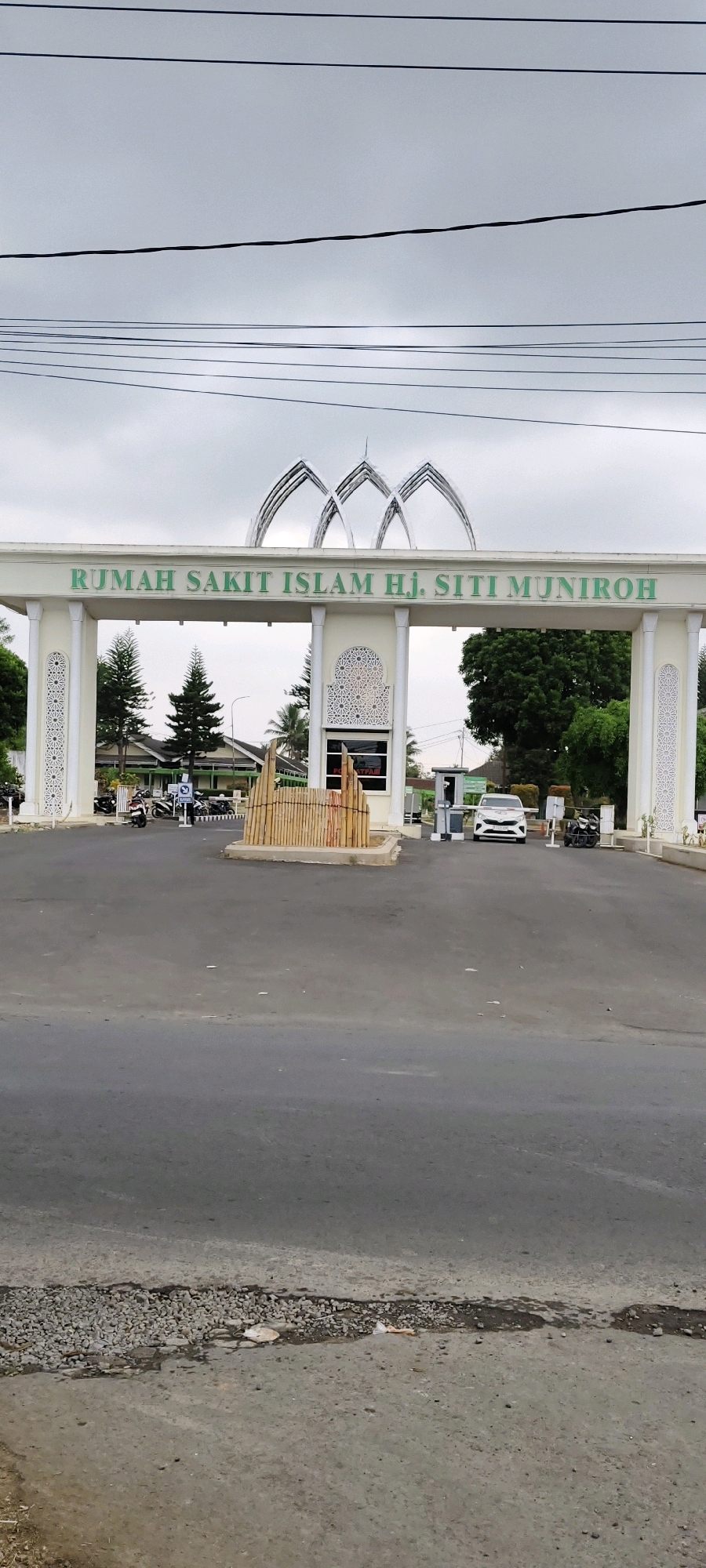 The image shows the main entrance of RUMAH SAKIT ISLAM HJ. SITI MUNIRROH in Tasikmalaya, Indonesia. The entrance features a large, ornate archway with the hospital's name written in green lettering across the top. The arch is supported by two pillars adorned with intricate white patterns. In the background, several cars and motorcycles are visible, suggesting the hospital is operational. A white sedan is positioned at the entrance, seemingly passing through a checkpoint or waiting to enter. The sky is overcast, and the overall lighting suggests a daytime scene. The road leading to the entrance shows some wear and tear, with patches of gravel and asphalt. There are also trees and buildings visible in the background, indicating a semi-urban or suburban environment.