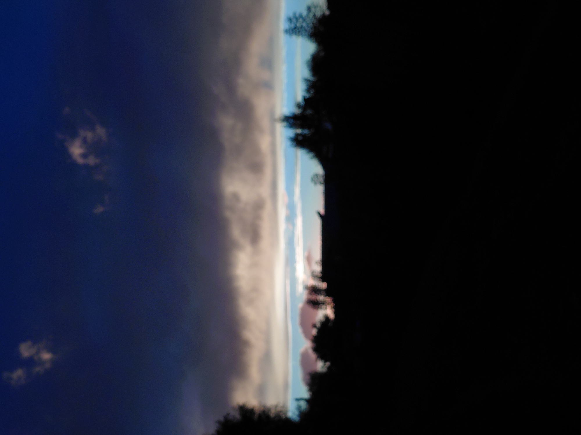 The image depicts a silhouetted landscape at dusk, with dark clouds dominating the upper portion of the frame. A band of lighter clouds, reflecting some residual light from the setting sun, stretches across the middle ground, transitioning to a pale blue sky. The foreground is entirely black, obscuring any ground-level details. A dark, indistinct line of trees and possibly buildings runs across the horizon, partially visible against the lighter sky. There are no visible people or animals. The overall impression is one of a tranquil, natural scene at the end of the day. Without any further context or identifiable landmarks, it is impossible to determine the specific location within Novosibirsk, Russia.