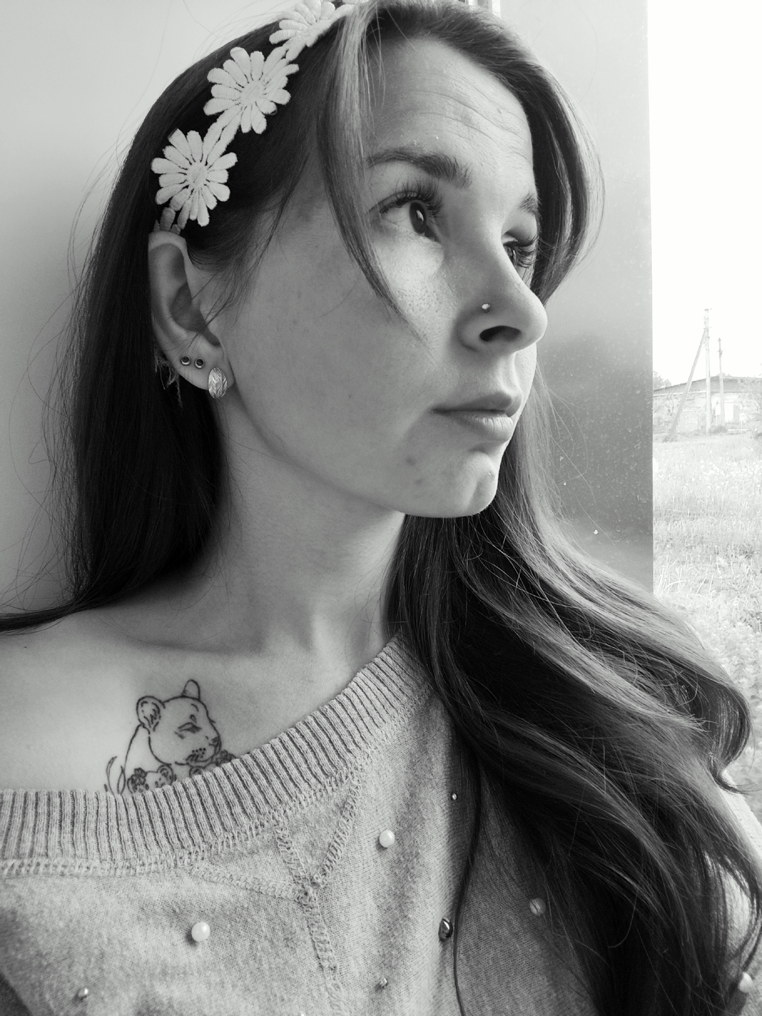 A grayscale photograph shows a woman looking out of a window. She has long wavy hair, a flower headband, and a lion tattoo on her collarbone. She is wearing a sweater adorned with pearls. The background through the window appears to be an overgrown field with a small building in the distance. There is no direct visual evidence in the image to confirm the location.