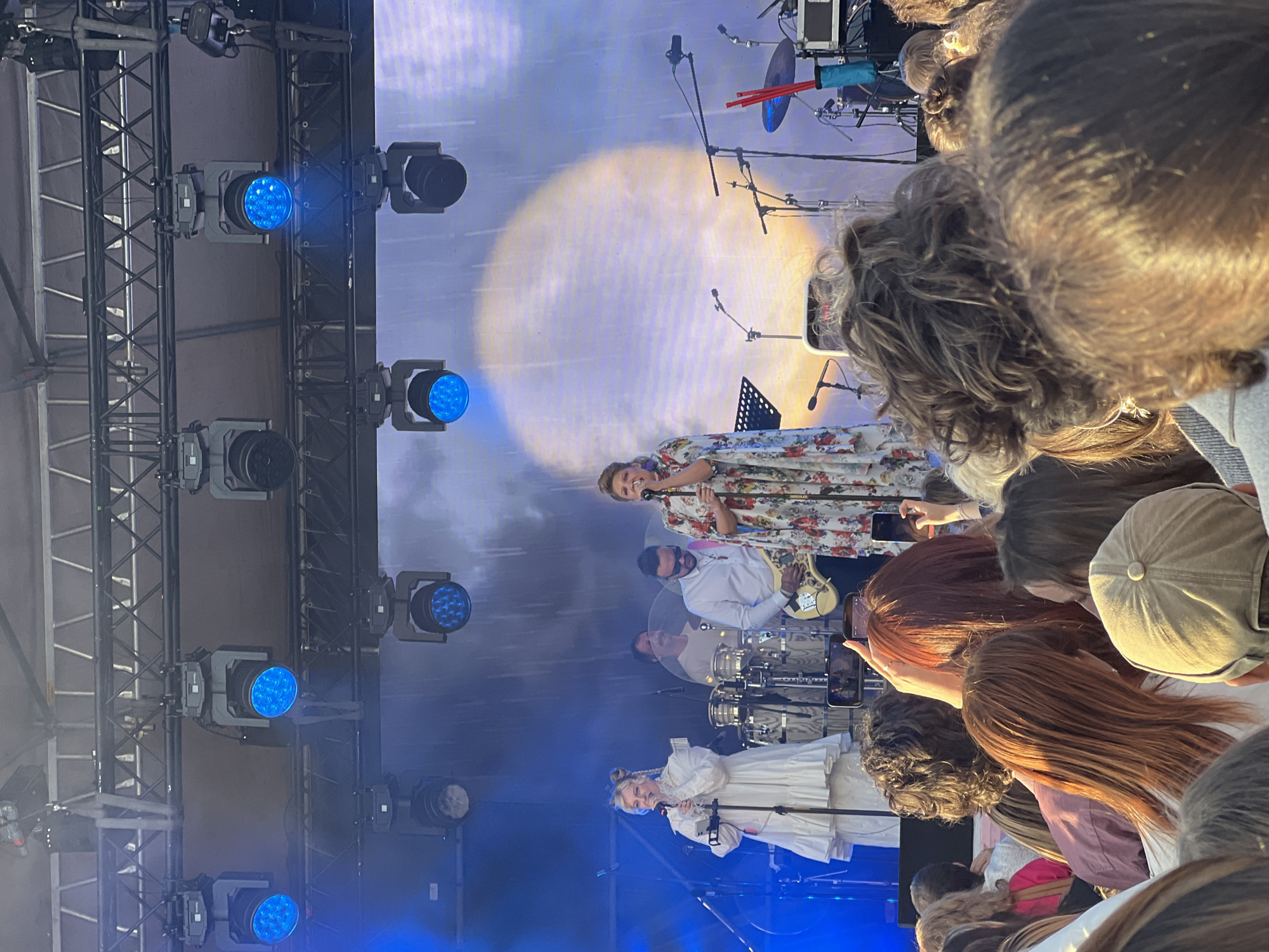 This image depicts an outdoor concert performance in Saint Petersburg, Russia. On stage, two female vocalists are prominent. The vocalist on the left, wearing a white ruffled dress, is singing into a microphone. To her right, another female vocalist, in a floral print dress, also sings into a microphone. Behind them, a male musician plays a guitar. A drum set and other musical instruments are visible, along with stage lighting and microphones. The backdrop features a large projection of a bright, circular light or moon. In the foreground, a crowd of concertgoers watches the performance. The overall atmosphere suggests a vibrant, live music event.