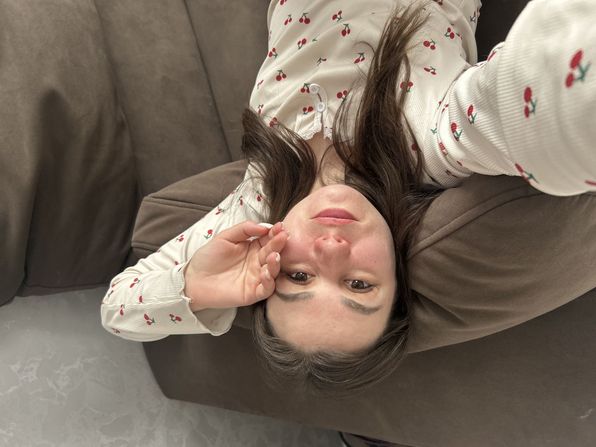 A woman with brown hair and brown eyes lies upside down on a brown sofa. She is wearing a light-colored long-sleeved top with a red cherry print. Her right hand is near her face. The background includes a dark brown sofa and a light-colored floor. The location is Chaikovsky, Russia.