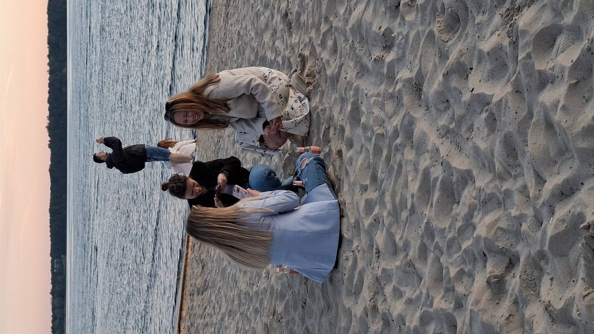 Three women are sitting on a sandy beach, near the water. One woman on the left is wearing a light blue blazer and ripped jeans. She is looking down at her phone. Another woman in the center is wearing a black jacket and blue jeans. She is also looking at her phone. The woman on the right is wearing a light-colored jacket and a floral dress. She is smiling and looking towards the left. Two bottles of pink liquid are visible on the sand near the woman on the left. In the background, a man is standing in the water and taking a photo with his phone. The sky is a soft pink and orange, suggesting it is sunset. The body of water appears to be a lake or a large bay. There are trees visible on the far shore. The scene appears to be a casual gathering on a beach. The image does not provide enough information to determine if it is located in Tallinn, Estonia.