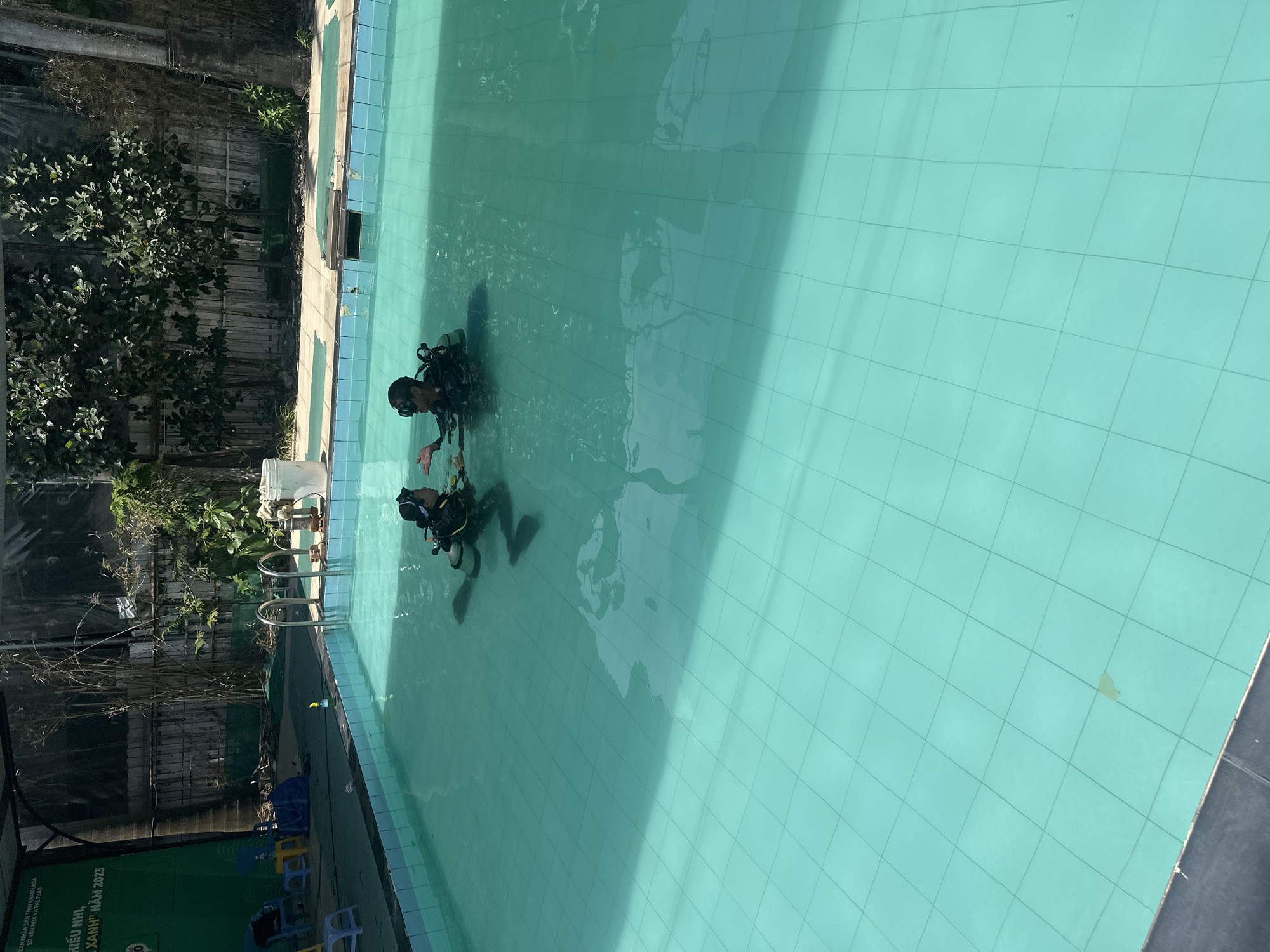 The image shows two individuals in scuba gear in a tiled, turquoise-colored swimming pool. They are positioned in the center of the pool, facing each other, seemingly engaged in some sort of underwater activity or training. The pool is surrounded by a deck on one side, and trees and greenery are visible beyond the deck. In the lower left corner of the image, there's a green banner, with some text that includes "Nha Trang", and a text of "2023". Based on this, it is likely that the location is within Nha Trang Ward, Vietnam. There are some blue and yellow chairs on the left side of the frame next to the banner.