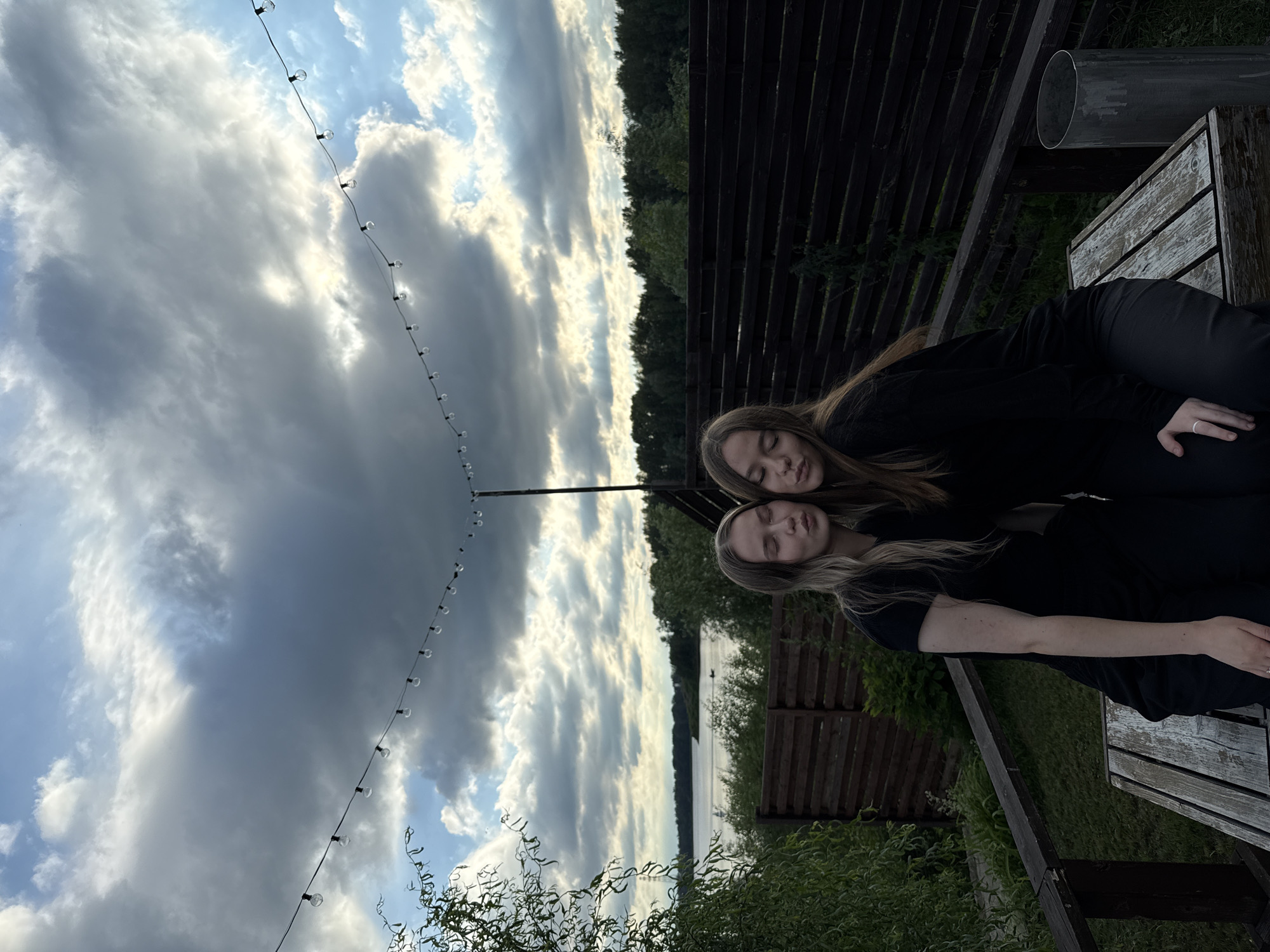 Two young women, appearing to be of Caucasian ethnicity, are posed together outdoors. One woman, on the left, has blonde hair and is wearing a black t-shirt. The other woman, on the right, has brown hair and is wearing a black long-sleeved shirt. Both women have their eyes closed and are puckering their lips, suggesting they are about to kiss or have just kissed. They are seated side-by-side on a weathered wooden bench. Behind them, a wooden fence runs horizontally, and beyond that, a body of water, likely the Kama River, is visible, with a tree line on the opposite shore. The sky above is dramatic, with a mix of dark, heavy clouds and patches of bright blue sky, with sunlight breaking through in rays. A string of globe lights is strung across the top of the frame, adding a decorative element to the scene. The overall impression is one of a casual, intimate moment in a natural setting, possibly a backyard or outdoor recreational area in Chaikovsky, Russia.