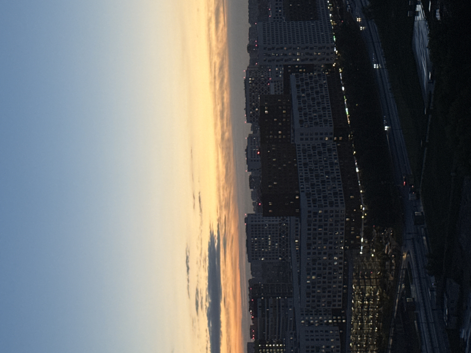 An aerial, high-angle, nighttime view captures a cityscape with apartment buildings and a highway in Dolgoprudny, Russia. The sky transitions from a deep blue at the top to a pale yellow and orange hue near the horizon, indicative of twilight or sunrise. Several multi-story residential buildings dominate the mid-ground and foreground, with windows illuminated by interior lights, creating a grid pattern. Some buildings have visible red lights on their roofs, possibly navigation beacons. A multi-lane highway runs through the lower portion of the image, with faint headlights from passing vehicles visible on the wet road surface, suggesting it may have rained or is raining. The overall scene is a typical urban landscape at dusk or dawn.