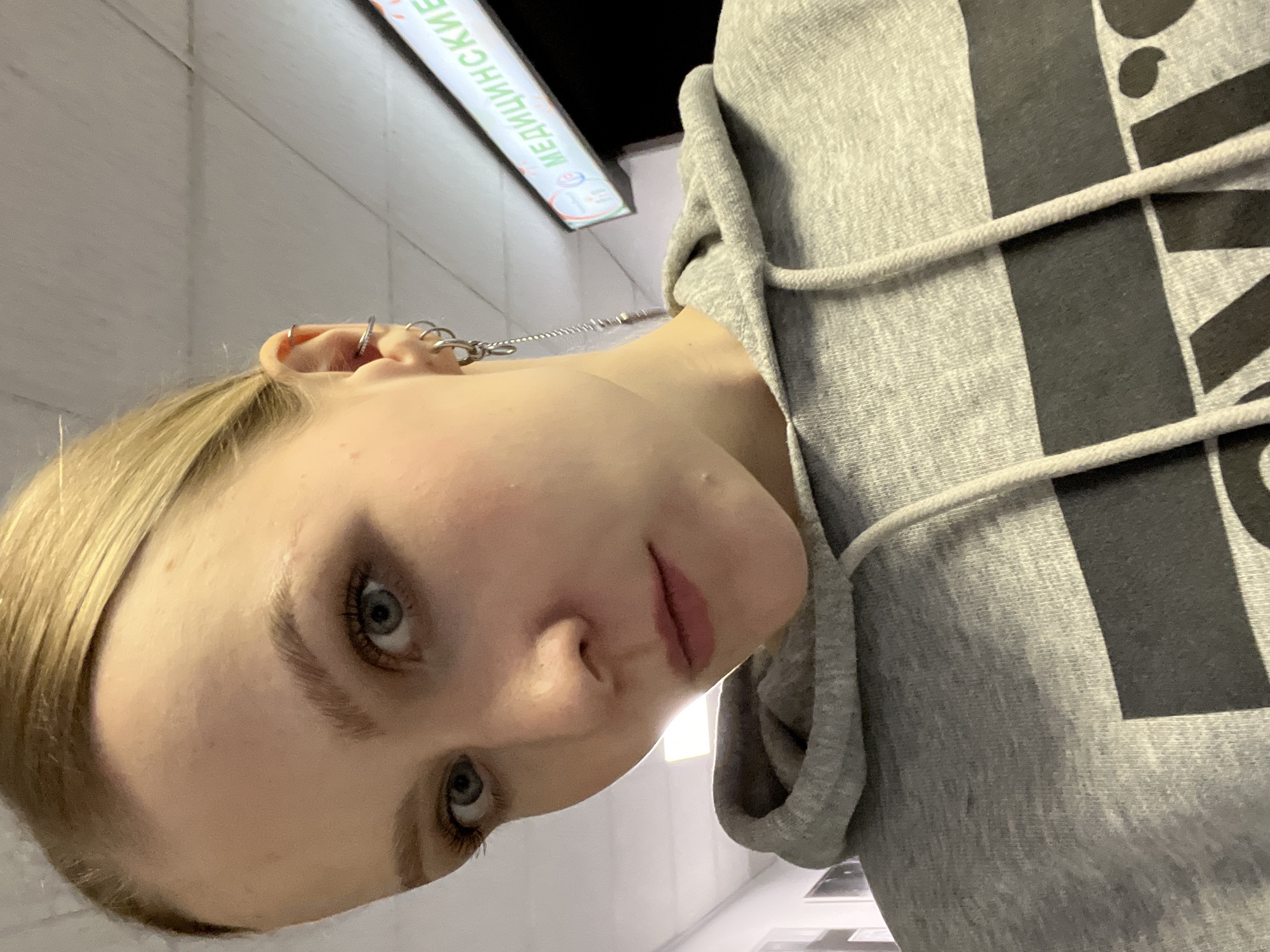 A young woman with blonde hair and multiple ear piercings wears a gray hoodie with a black design. She is looking down at the camera. The background shows a sign in Russian, "МЕДИЦИНСКИЕ" ("Medical"), suggesting the location is in the Khimki area of Russia. The image is taken from a low angle.