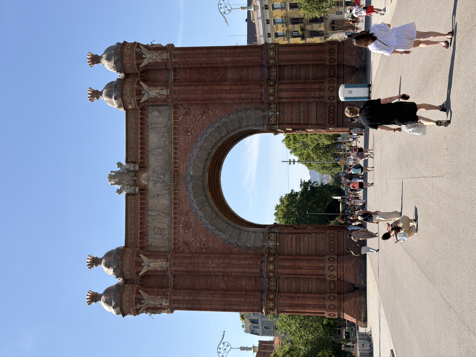 The image depicts the Arc de Triomf in Barcelona, Spain, a triumphal arch constructed from red brick. The arch is grand and ornate, with decorative carvings and sculptures adorning its structure. It is situated at the entrance to the Parc de la Ciutadella.
In the foreground, the paved road leads towards the arch, with several people walking. A woman in a white dress walks away from the camera, and a man in a black t-shirt and shorts walks ahead of her, carrying a white shopping bag. Another person, dressed in beige, is crouched down on the road, possibly taking a photo or adjusting something. Further back, a variety of people are seen walking and milling about, some in groups, suggesting a lively atmosphere.
The background shows more of the park, with trees lining the path leading through the arch. Buildings are visible in the distance, with balconies and architectural details characteristic of Barcelona. The sky is clear and blue, indicating a sunny day. A portable toilet is visible on the right side of the road. The scene is busy, with tourists and locals enjoying the public space.