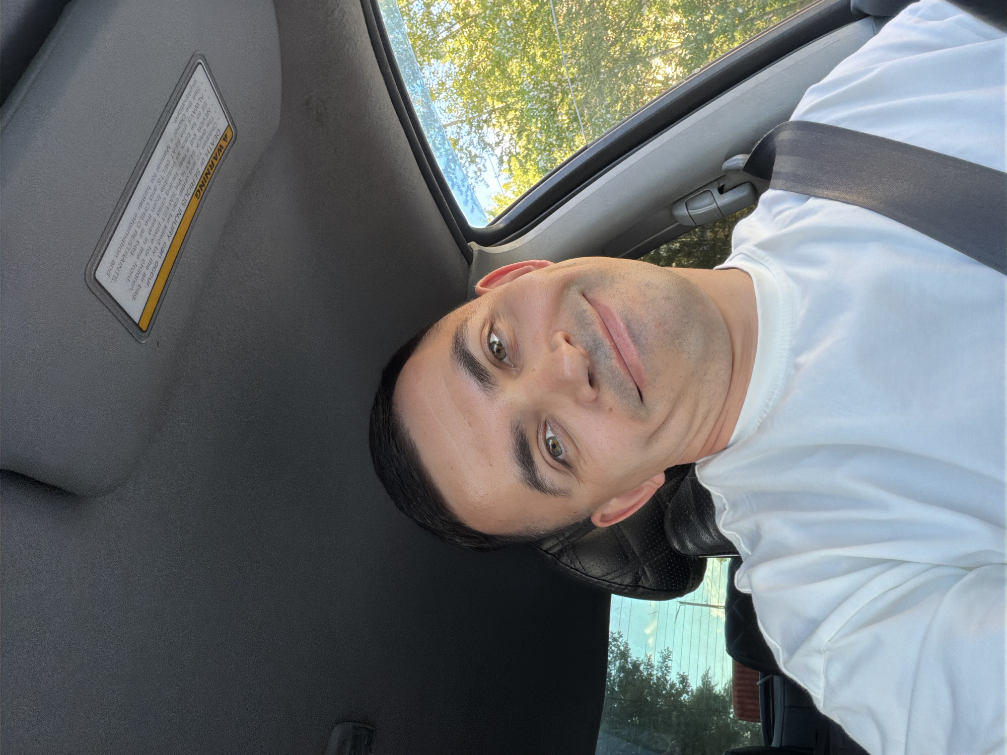 A rotated selfie captures a man inside a car during daytime. The subject, an adult male with short dark hair, dark eyebrows, and striking light-colored eyes, looks directly at the camera with a subtle, confident smile. He wears a plain white short-sleeved t-shirt, and a dark seatbelt is fastened across his chest, suggesting he is in the driver's or front passenger seat.
The car's interior is visible, including a grey sun visor on the left. Affixed to the visor is a prominent "A WARNING" label with black text on a white background, framed by a yellow border. The text reads: "DEATH OR SERIOUS INJURY can occur. The BACK SEAT is the SAFEST place for children. Children 12 and under can be killed by the air bag. NEVER put a rear-facing child seat in the front. Sit as far back as possible from the air bag. ALWAYS use SEAT BELTS and CHILD RESTRAINTS. See the Owner's Manual for further information and explanations."
Behind the man's head, a black, possibly leather, car seat or headrest is partially visible. Beyond the car's windows and sunroof, bright natural light filters through, illuminating lush green foliage from trees and a clear sky, indicating sunny weather. The scene appears to be set outdoors in Izhevsk, Russia.