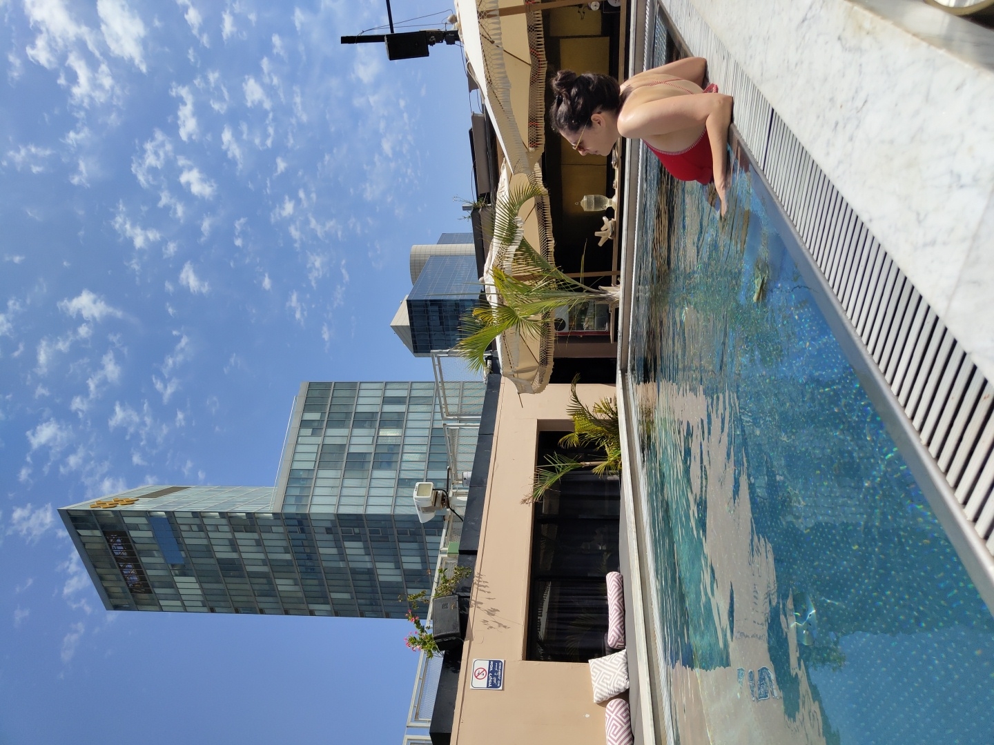 This image depicts a vibrant rooftop swimming pool scene with a city skyline under a bright blue sky. A woman, with dark hair tied back in a bun and wearing sunglasses, is visible in a red swimsuit, leaning over the edge of the pool and looking intently towards the distant cityscape. The pool water is clear, revealing a shimmering blue mosaic pattern on the bottom, and is framed by white marble coping.
In the background, several modern high-rise buildings with glass facades rise against a bright blue sky dotted with scattered fluffy white clouds. One prominent building features a yellow Pelephone logo on its upper section. Palm trees and other greenery, along with partial views of white umbrellas and patterned cushions, surround the pool area, suggesting an outdoor lounge setting. A black antenna-like structure is visible against the sky on the far right.
The scene conveys a bright, sunny daytime in an urban environment, identified as Tel-Aviv, Israel. A white sign with a no-smoking symbol and the Hebrew text "אסור לעשן" (No Smoking) is visible on a black speaker near the pool edge. The overall atmosphere is one of relaxed enjoyment amidst an urban backdrop.