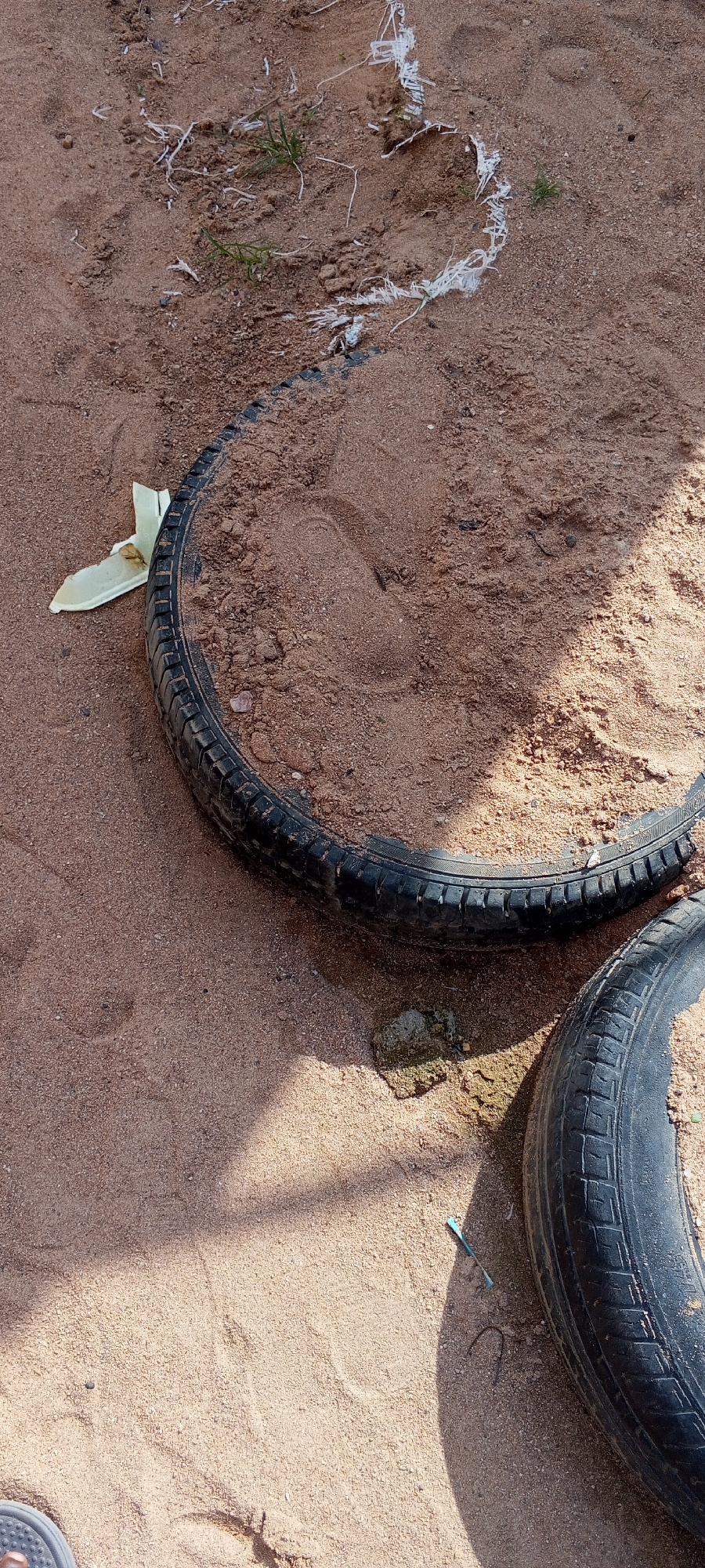 Two bicycle tires are partially buried in sand. The tires are black and have visible tread patterns. One tire is positioned in the upper left quadrant of the image, filled with sand, and is slightly angled. The other tire is in the lower right quadrant, also filled with sand. The sand is light brown and appears to be a mixture of fine grains and small pebbles. There are a few blades of grass growing out of the sand near the upper tire. Some white, fibrous material, possibly plastic debris, is scattered on the sand in the upper portion of the image. A small, light-colored piece of debris, possibly plastic, is near the edge of the upper tire. The scene suggests an outdoor environment, possibly a yard or public space in Jalingo, Nigeria, where the tires have been discarded or left. The lighting indicates it is daytime, with shadows cast on the sand.