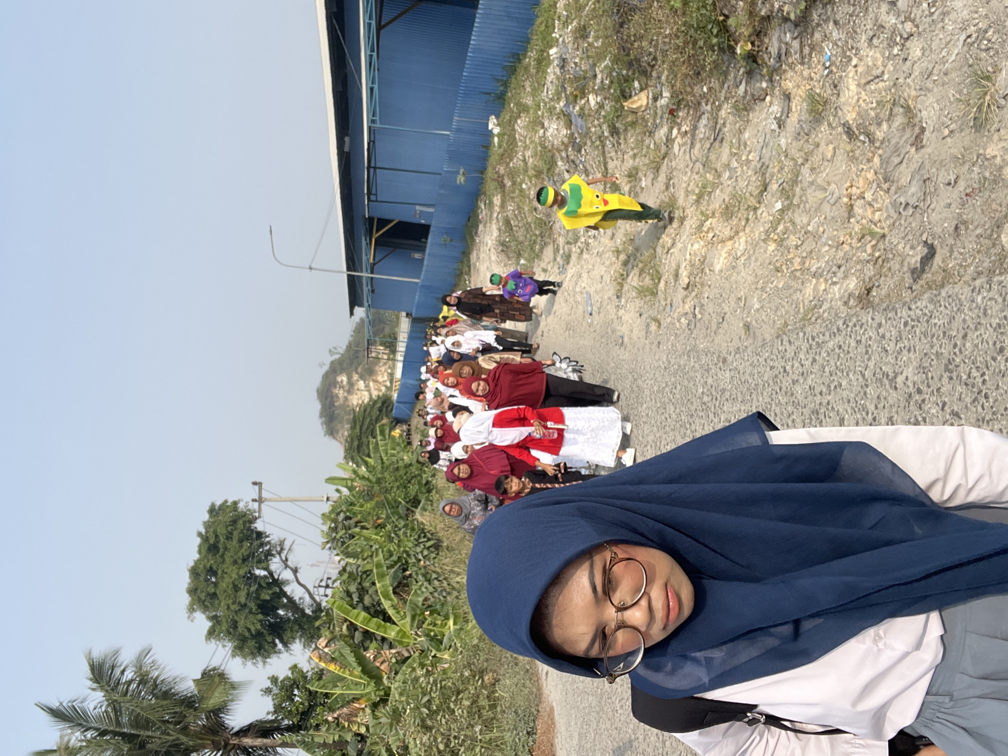 In Klapanunggal, Indonesia, a group of people, primarily women and children, are walking along a dirt road on a sunny day. The group is diverse, with some women wearing hijabs and modest clothing in various colors, while some children are dressed in costumes. The foremost person in the image is a young woman wearing a dark blue hijab and eyeglasses, looking downwards. Behind her, the procession continues with a mix of adults and children. The environment is characterized by lush green vegetation, including banana trees, and a clear blue sky. In the background, a long, blue industrial building with a white roof is visible, along with a power pole and some rocky terrain. The road itself is a rough, unpaved surface.