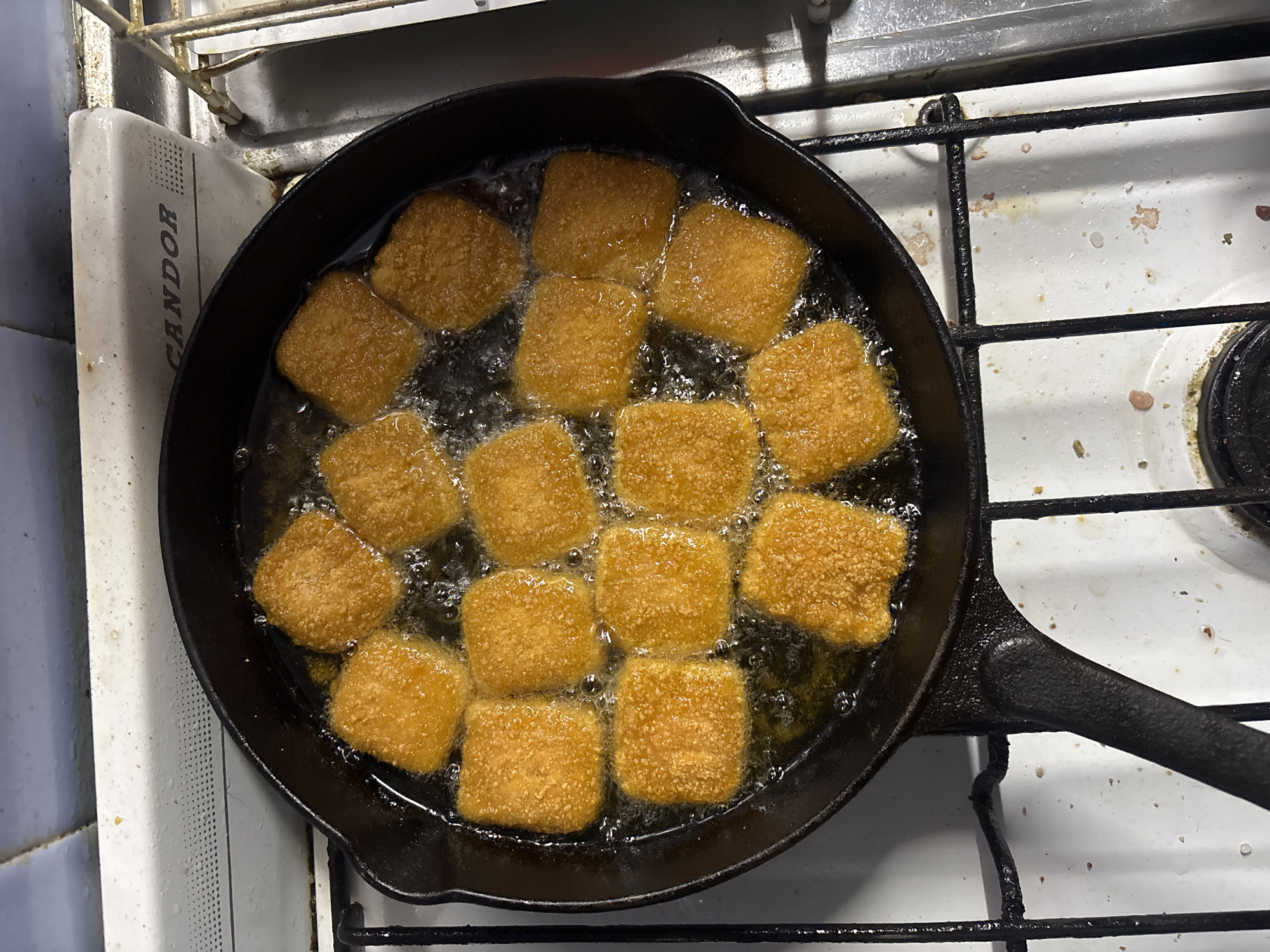 A cast iron skillet containing approximately 14 square, breaded items is sitting on a white gas stove top. The items are submerged in oil and appear to be frying. The stove is adjacent to a white cabinet with the word "CANDOR" printed on it. The location is likely a kitchen in Buenos Aires, Argentina based on the brand of the appliance.