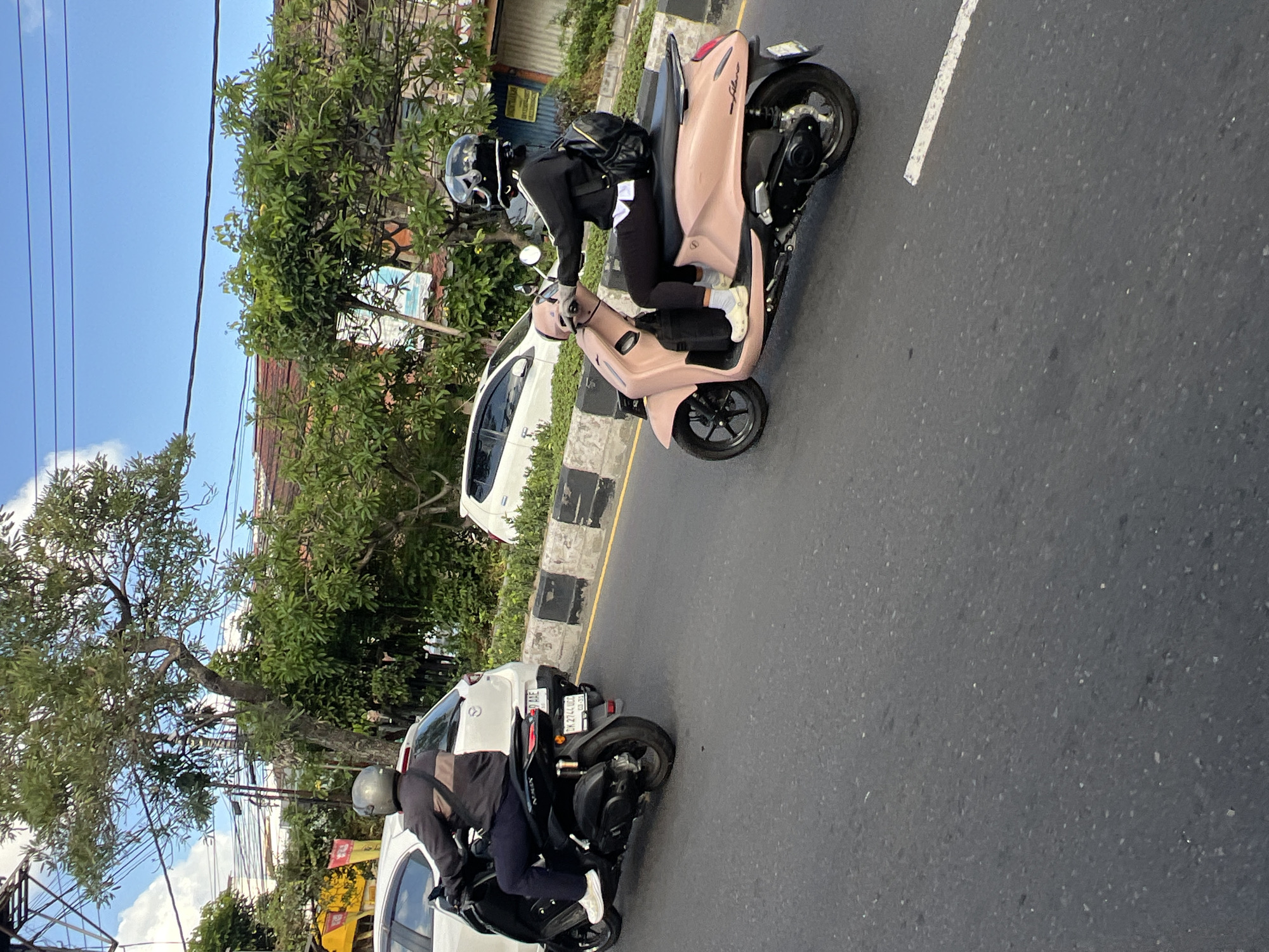 Two individuals on scooters are traveling along a street in Kuta, Indonesia. The person in the foreground is riding a pink scooter, wearing a black helmet with a white design, a black jacket over a white shirt, black leggings, and white crocs. They have a black backpack. The person in the background is on a black scooter, wearing a grey helmet, a dark jacket with a tan stripe on the shoulder, and dark pants. They are behind a white car. The scene is characterized by a paved road, a concrete barrier with black and white markings, lush green trees, and buildings in the background under a bright blue sky with scattered clouds. Utility lines are visible overhead. The license plate of the black scooter is visible and reads "DK 2744 UQA".