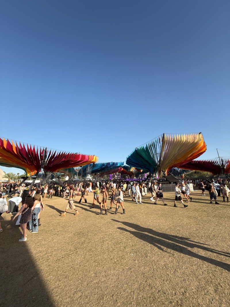 This full shot captures an outdoor music festival or large event under a clear, bright blue sky in Indio, United States. The scene is dominated by a wide, open field covered in dry, yellowish-brown grass, characteristic of an arid climate. Numerous people are scattered across the field, walking in various directions. Many individuals are dressed in diverse festival attire, ranging from casual to more elaborate or revealing outfits, including boots and hats.
Prominent in the midground are several large, artistic installations. These structures feature flowing, draped fabrics or panels in a spectrum of vibrant rainbow colors, creating a fanned or layered effect. These colorful art pieces extend horizontally across the frame, contributing to the festive atmosphere. In the background, more people and event infrastructure are visible, indicating the scale of the gathering.
The time of day appears to be late afternoon, as evidenced by the long shadows cast by the people and the colorful structures stretching across the grassy ground from left to right. The clear sky suggests sunny and pleasant weather conditions for the outdoor event. There is no visible text in the image. The overall impression is one of a lively, bustling festival setting.