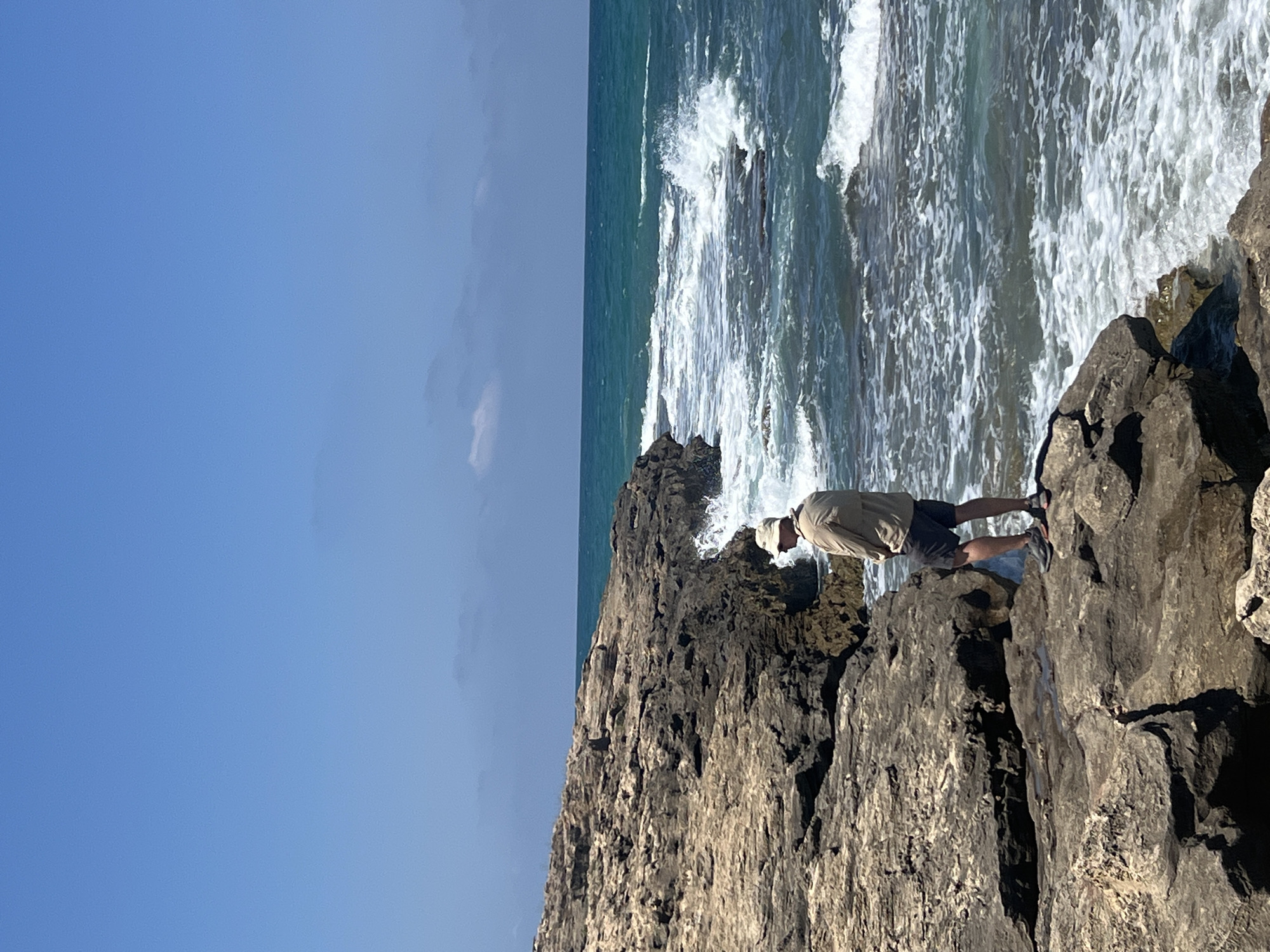 A man stands on a rocky coastline in Hof HaCarmel Regional Council, Israel, facing the sea. He is wearing a light-colored long-sleeved shirt, dark shorts, a hat, and athletic shoes. The man is positioned on a dark, textured rock formation, with the blue ocean stretching out before him. Waves crash against the rocks, creating white foam. The sky above is a clear, bright blue with a few wispy clouds. The environment is rugged and natural, characteristic of a coastal landscape.