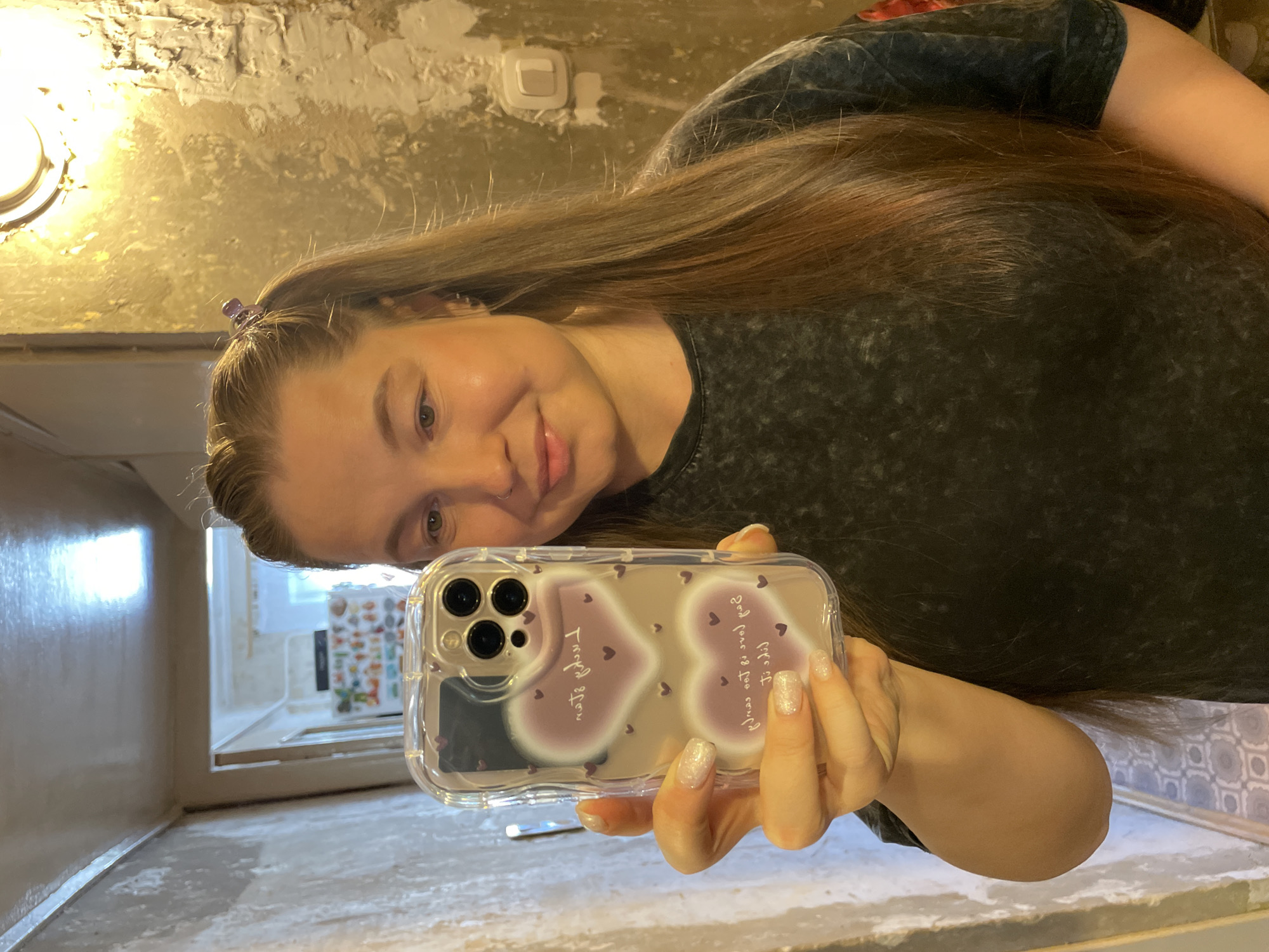 A mirror selfie captures a woman indoors in Izhevsk, Russia, with a pleasant, soft smile. She has long, straight brown hair, with a small purple claw clip securing some strands on the right side of her head. She is wearing a dark, possibly charcoal or mottled grey, t-shirt.
In her right hand, she holds a clear-cased iPhone (likely a Pro model with three lenses), which is decorated with two large, faded pink-purple hearts and numerous smaller dark hearts. The text on the phone case, when read correctly (un-mirrored), says: "I hope your life is filled with peace" on the heart closer to the phone's top, and "I hope your love is filled with joy" on the heart closer to the bottom. Her fingernails are painted with a shimmery, light-colored polish.
The background reveals an informal interior setting. A rough, textured wall, possibly unfinished plaster or concrete, is painted in a golden or yellowish hue, with some areas appearing darker or less finished. A round, warm-toned light fixture is mounted on the wall to the upper left, providing artificial illumination. A white electrical switch is also visible on the wall. The surface below the mirror appears to be a light-colored, rough material like concrete or stone. Through a reflection in the background, another room can be seen with a window and what appear to be shelves holding various items. The lighting suggests it is either evening or an indoor space heavily reliant on artificial light, and the overall environment hints at a functional or utilitarian space, possibly undergoing renovation.